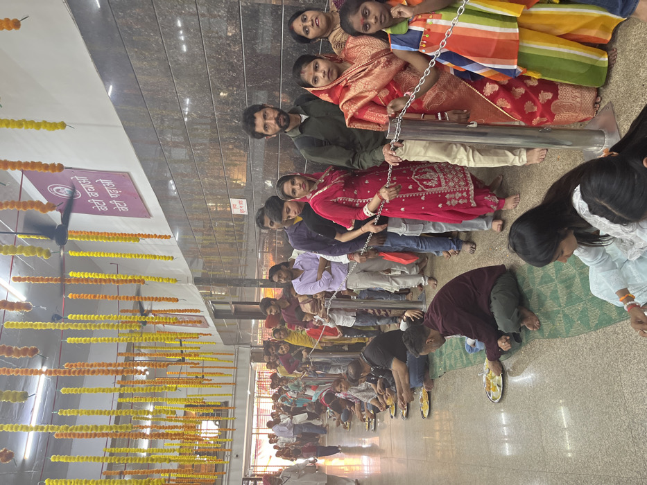 The image depicts a scene in a temple in Panchkula, India, located at the coordinates 30.723 latitude and 76.862 longitude. The scene shows a line of people waiting to receive food. The people are dressed in a variety of clothes, and many are wearing traditional Indian attire. There are several metal poles with a chain strung between them, forming a barrier. The temple walls are covered with decorative tile and a sign hanging on the wall that reads "Beti Bachao Beti Padhao". There are also yellow and orange floral garlands hanging from the ceiling. The floor is tiled and there are green mats on the floor where people are sitting. The people in the line are interacting with each other and some appear to be speaking. The lighting in the scene is soft and warm, and there is a feeling of peace and tranquility.