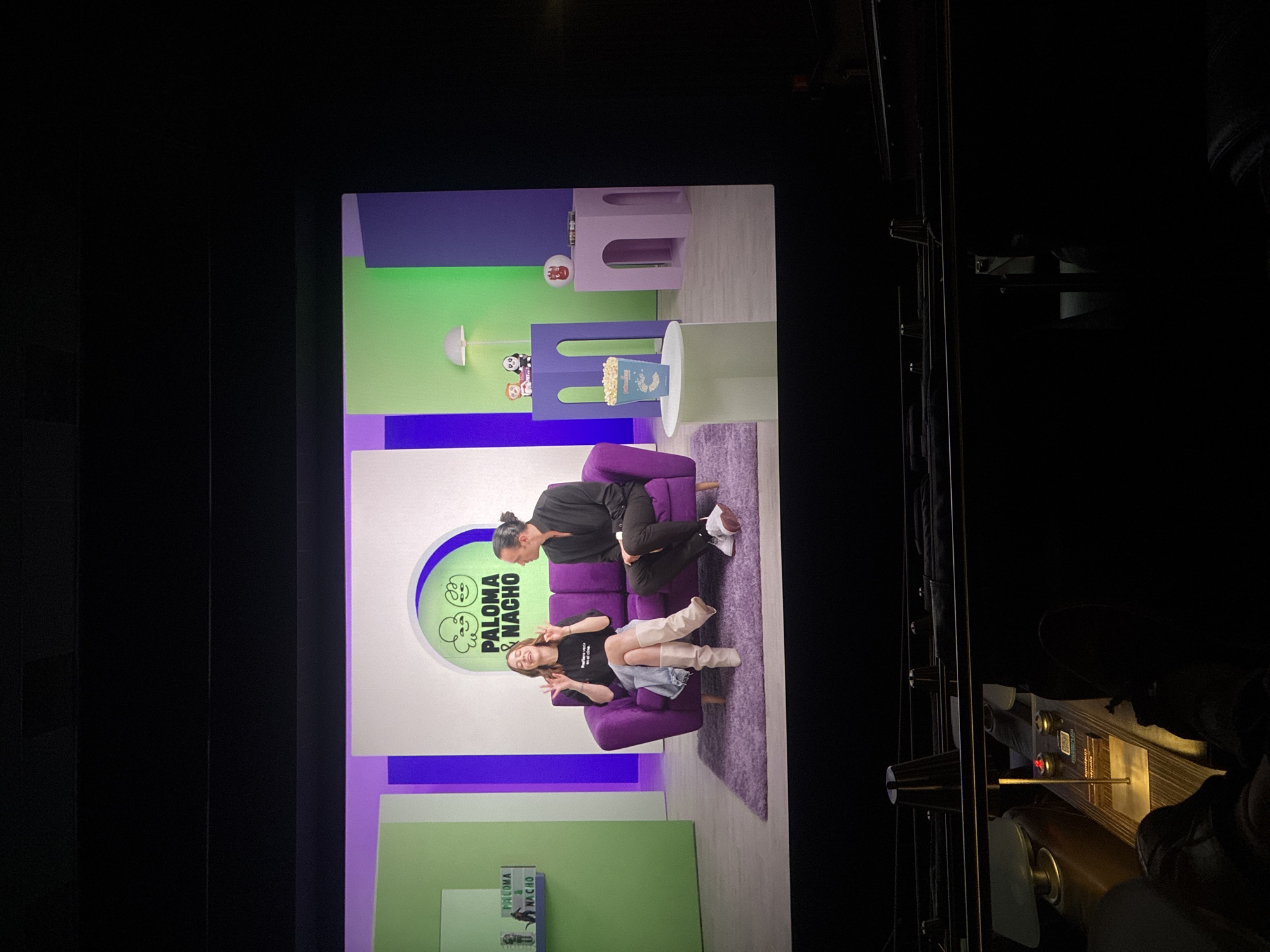The photograph was taken inside a movie theater at Cinepolis Miyana in Mexico City. It shows a movie screen displaying a set with two people on a purple couch. The set has green and purple walls. A table next to the couch has a box of popcorn. The theater seating and equipment are visible in the foreground.