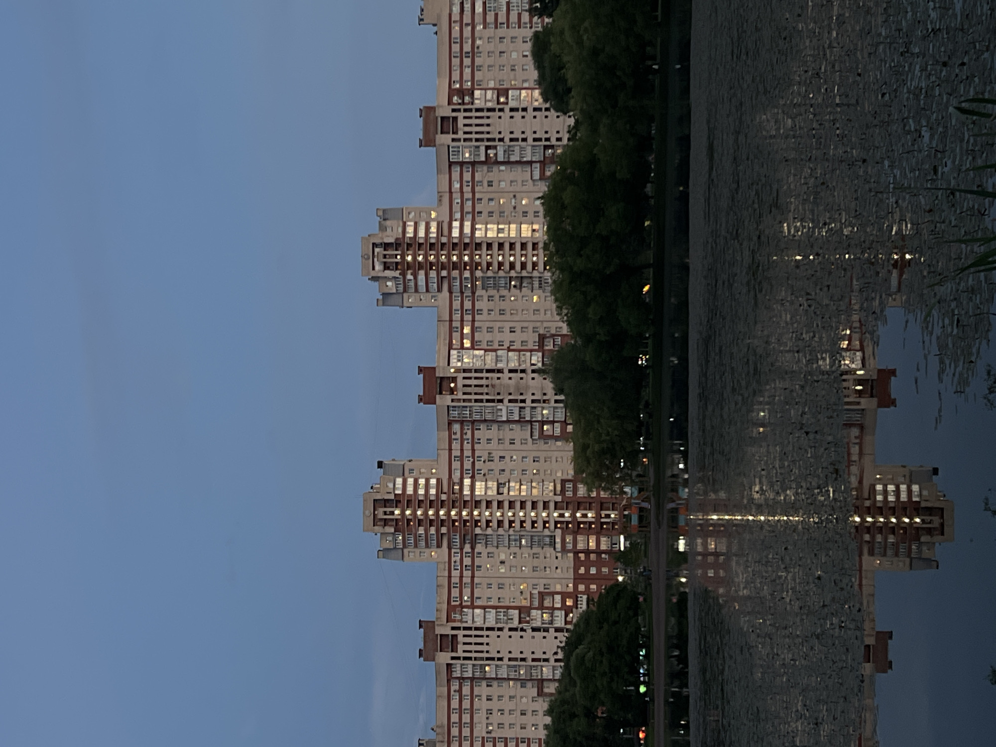 The image depicts a tall, multi-story residential building with a light-colored facade and red accents, reflecting in the calm water of a body of water. The building has numerous windows, many of which are illuminated, suggesting it is evening or night. Trees line the edge of the water and the building, with some greenery visible in the foreground. The sky is a clear, pale blue, indicating a twilight setting. The reflection of the building in the water is distorted by ripples, creating an abstract pattern. The scene appears to be a tranquil urban setting, possibly a park or residential area with a waterfront. There is no visible text in the image.