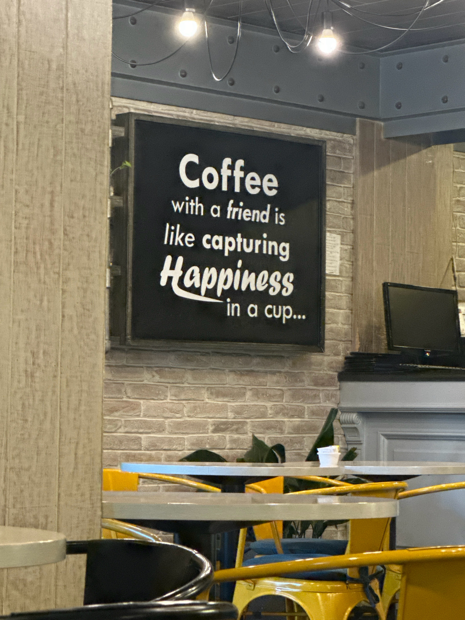The image depicts the interior of a coffee shop. Dominating the center is a black sign with white lettering that reads "Coffee with a friend is like capturing Happiness in a cup...". The sign is mounted on a brick wall. To the left of the sign, a vertical wooden panel is visible. In the background, there is a computer monitor on a counter. Several tables and chairs, some with bright yellow frames, are arranged in the foreground and mid-ground. The lighting appears to be from overhead fixtures, one of which is clearly visible. The overall ambiance suggests a casual and welcoming environment. This location is likely in Kathmandu Metropolitan City, Nepal, as specified by the user prompt, though the image itself provides no definitive visual cues to confirm this specific geographical location.