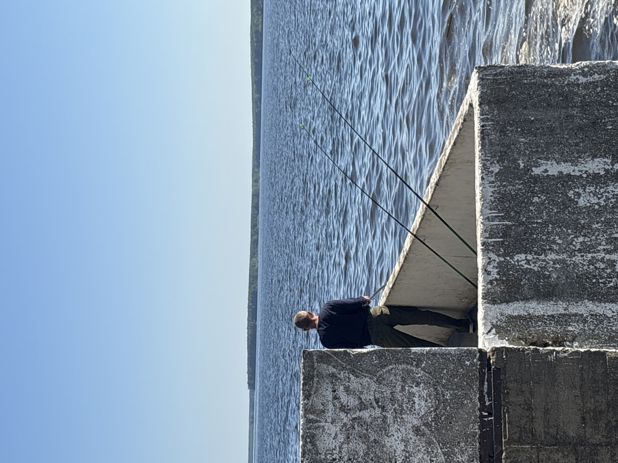 A man is fishing on a concrete structure that protrudes into a body of water. The location is likely the Kama River, Chaikovsky, Russia. The man, wearing a dark shirt and pants, is holding fishing lines. Three fishing rods are positioned at the edge of the structure extending into the water. The water surface is rippled. The sky is clear and blue. The concrete structure appears aged and weathered. A line of trees is visible in the distance.