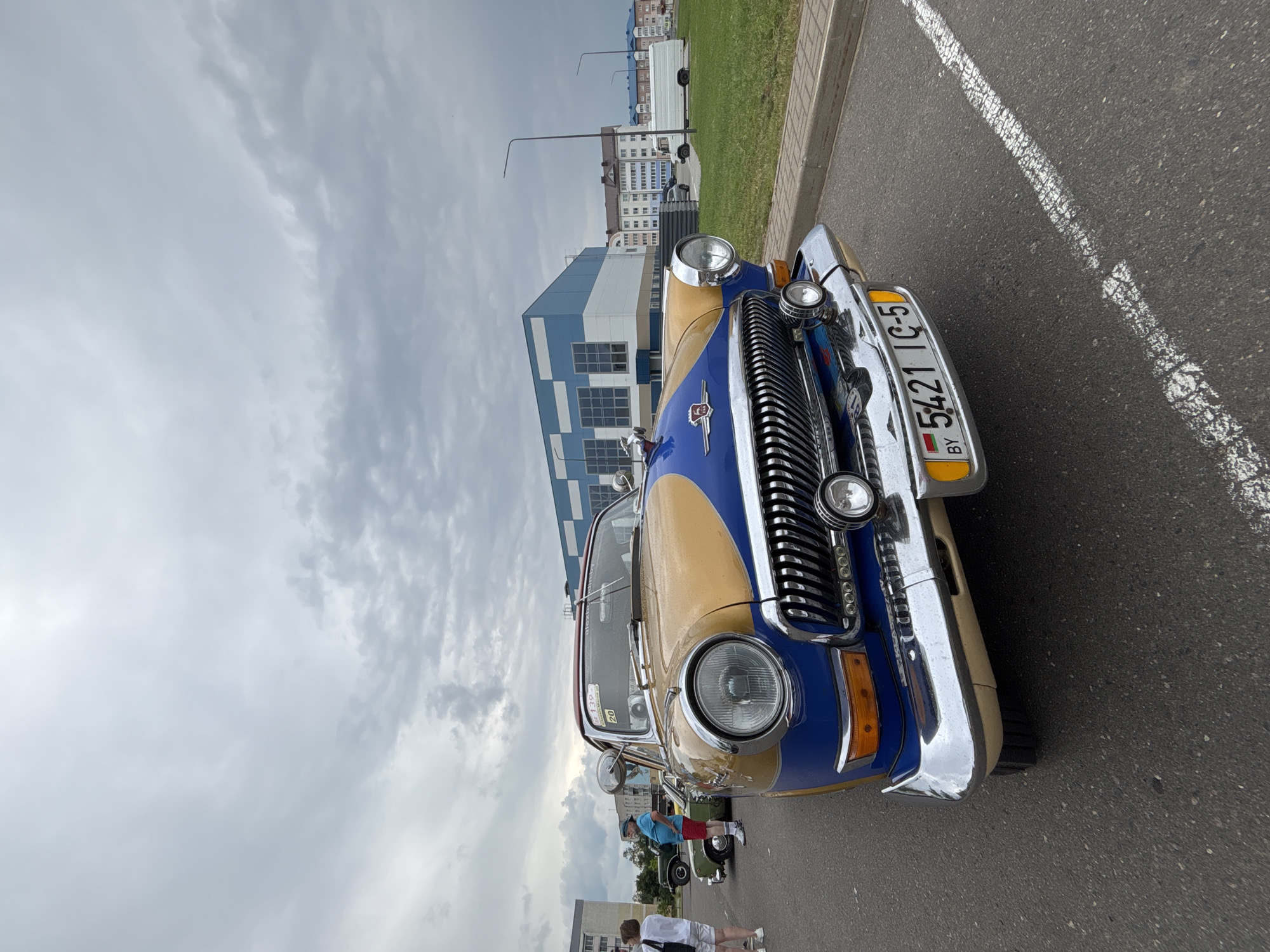 In the city of Uzda, Belarus, an aged, two-toned car, yellow and blue, is parked on asphalt. The car's chrome grille and circular headlights gleam under an overcast sky. A license plate displaying "BY 5421 IC-5" is affixed to the front. In the background, a modern building with blue and white accents stands, along with residential buildings and a white truck. Several individuals are visible near other vehicles, one wearing a blue shirt and red shorts, and another a white shirt and shorts. The scene suggests an outdoor event or gathering focused on vintage automobiles.