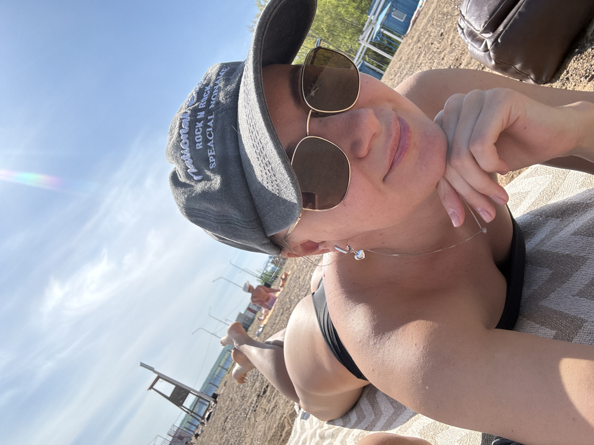 A woman lies on a towel on a beach, wearing a black bikini and a gray baseball cap with text. She is also wearing sunglasses and a necklace with a heart-shaped pendant. Her head is resting on her hand, and she is smiling. Behind her are other people on the beach, as well as wooden structures and a blue building. The photograph was taken at a beach in Chaikovsky, Russia.