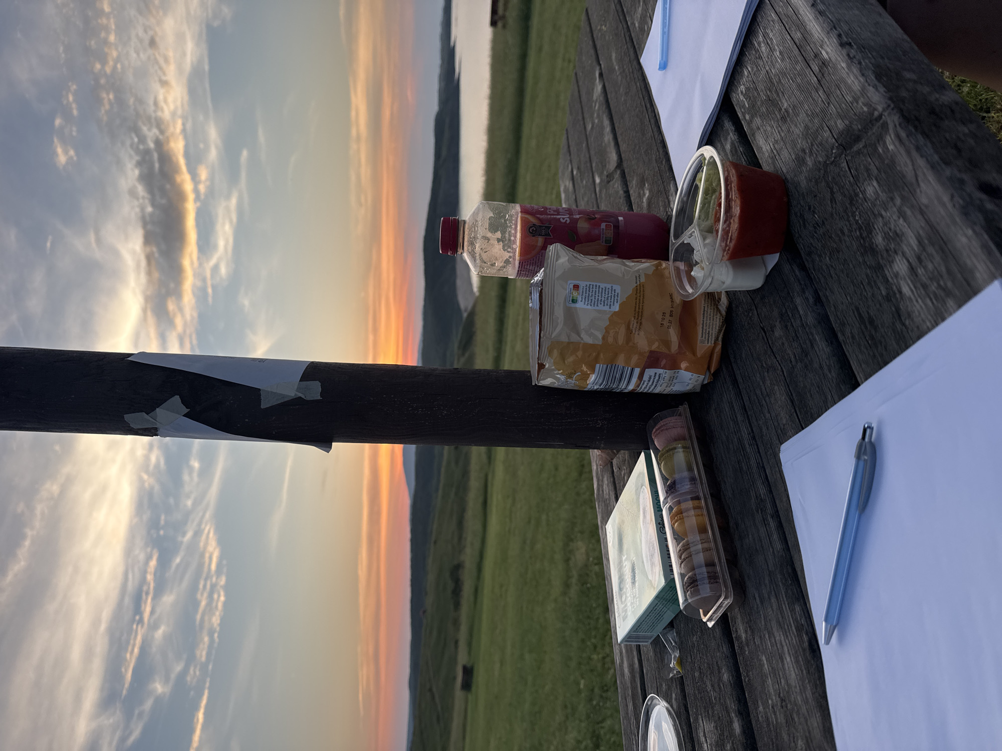 The image displays an outdoor scene at sunset, situated on a wooden picnic table with a panoramic view of the landscape of Sinemorets, Bulgaria.
In the foreground, a dark, weathered wooden picnic table occupies the lower portion of the frame. Arranged on the table are several items: a partially open bag of chips with orange and yellow packaging stands to the left of a central wooden post. To its right, a clear plastic bottle with a red cap contains a reddish-pink liquid, labeled "Peach Superfruit." A transparent plastic container holding six distinctively colored macarons (brown, orange, yellow, green, purple, and pink) is positioned next to a light green box labeled "Mochis Coconut." A clear plastic cup with two internal compartments, one containing a white creamy substance and the other a red sauce, is visible in the lower right. Two stacks of white paper are on the table; one in the lower left has a silver-blue pen resting on it, and another partially visible stack in the lower right also has a blue pen. A small clear plastic dish and a crumpled yellow wrapper are also present.
A prominent, dark brown wooden post rises vertically through the center of the image, from the table level into the sky. Several pieces of white paper are attached to this post with light-colored adhesive tape.
The background reveals an expansive landscape under a sunset sky. Verdant rolling hills and fields stretch across the midground. A rectangular structure, possibly a hay bale, is visible in the mid-distance within the fields on the left. In the further distance, particularly to the right, a body of water, likely the Black Sea or a large lake, is visible, bordered by undulating landforms. The horizon features faint outlines of distant mountains or hills. The sky transitions from vibrant orange and red hues near the horizon, characteristic of a sunset, to lighter yellows and blues higher up, with scattered wispy and stratified clouds illuminated by the low sun. No individuals are visible in the scene.