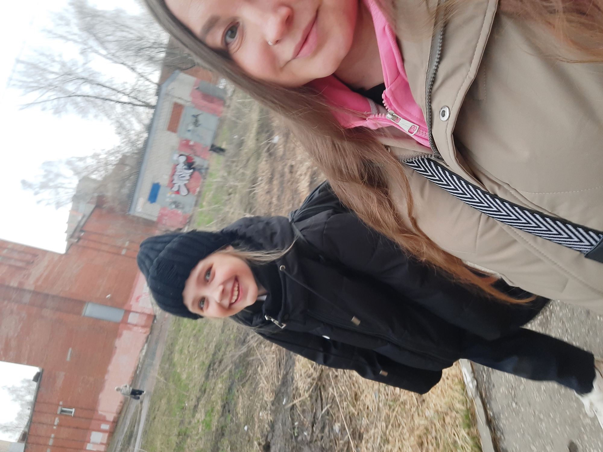 The image depicts two females in an outdoor environment, likely in the city of Izhevsk, Russia, based on the architectural style and overall aesthetic. The first female, likely a young adult, is taking the selfie. She has light skin, long brown hair, and is wearing a beige jacket with a pink hoodie underneath. She has a nose piercing on the left side. The second female, likely a child, is wearing a black jacket and a black knit cap. She is smiling at the camera.
The background reveals a somewhat urban environment. Several brick buildings are visible, one of which has graffiti on its side. The ground appears to be a mix of grass, dirt, and possibly pavement. A tree is present, indicating an outdoor location. Another person is walking in the background.