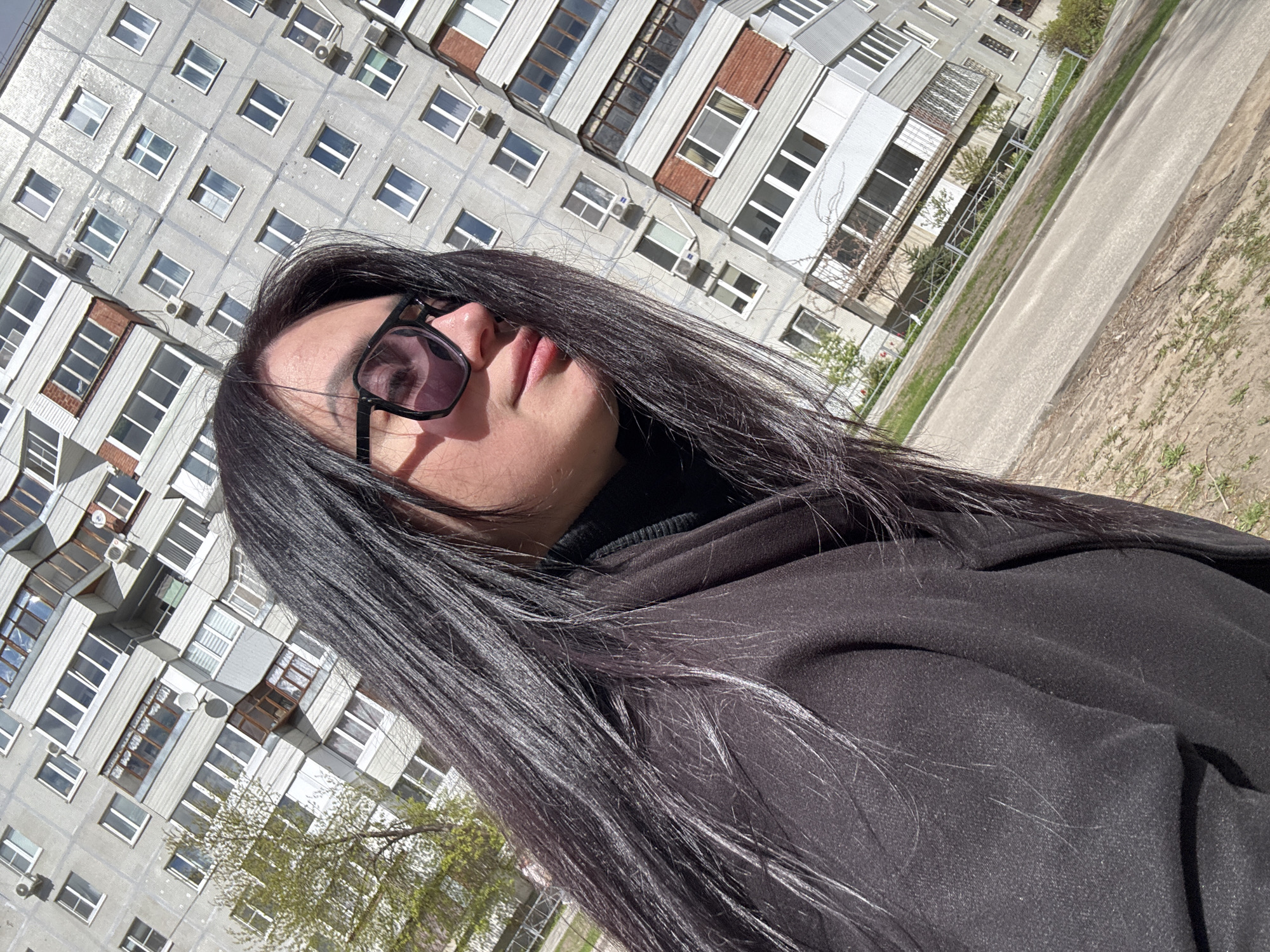 The image features a woman posing outdoors on a sunny day. She is seen from the chest up, looking slightly upwards and to her left, with her head tilted. She has long, straight, dark hair, which is illuminated by bright sunlight, and is wearing black-framed sunglasses with dark lenses. Her attire consists of a black coat or jacket worn over a black turtleneck.
In the background, a large, multi-story building with a light grey facade dominates the scene. This building features numerous rectangular windows, some with white frames, and several air conditioning units mounted on its exterior. The architecture suggests a functional, possibly Eastern European or post-Soviet, residential or office building.
To the right of the woman and in the mid-ground, a paved path or road is visible, bordered by sparse patches of green grass and dry earth. Some faint green foliage from distant trees or bushes can be discerned near the building. The entire photograph is captured at a significant diagonal angle, giving the scene a dynamic and unconventional perspective. The strong sunlight indicates it is daytime, and the weather appears clear and bright. No discernible text is visible in the image. The setting appears to be an urban or suburban environment.
