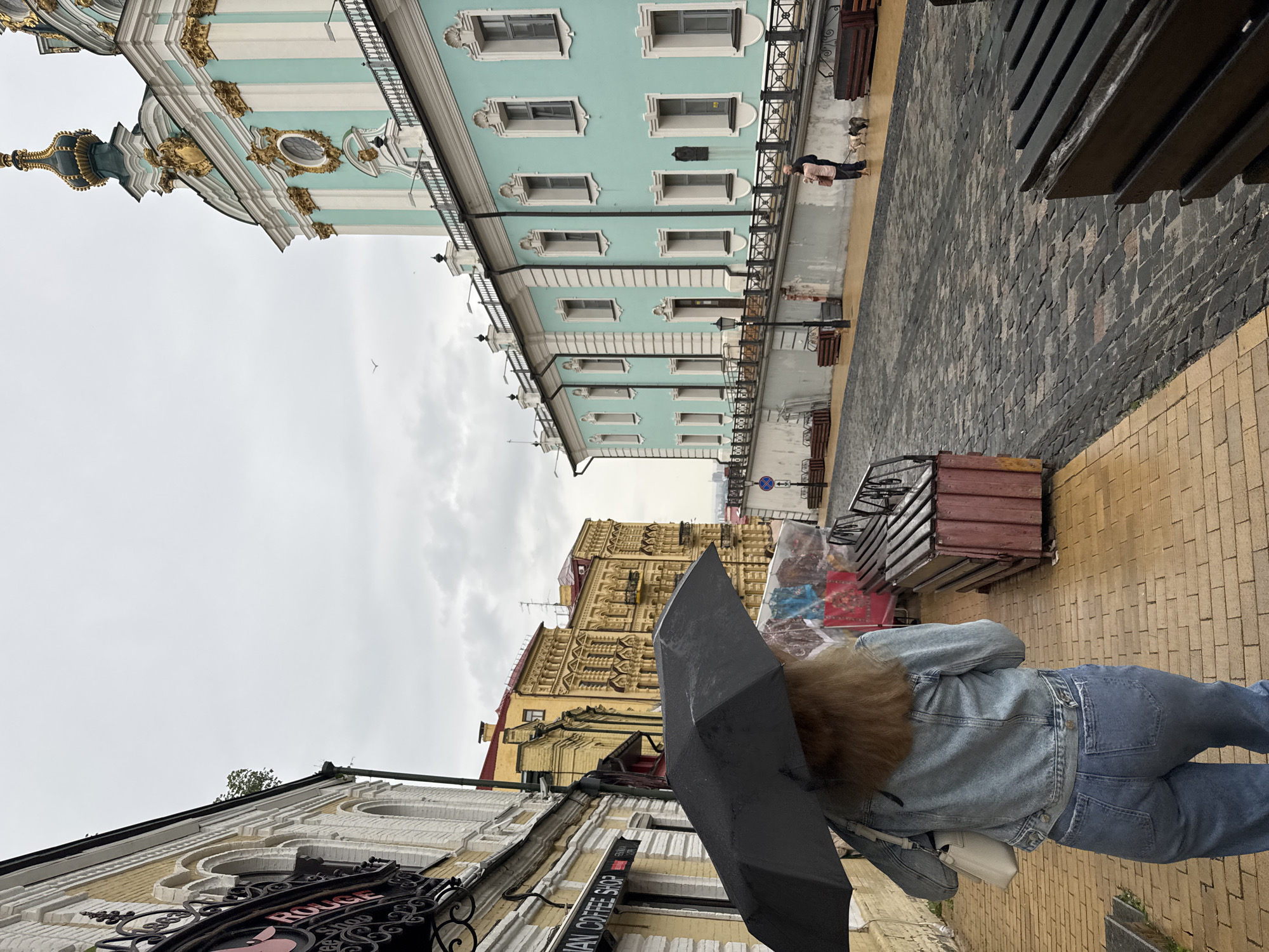 This media file shows a street scene in Kyiv, Ukraine, under an overcast sky. The image is taken from a low angle, looking up a street lined with historic buildings.
In the foreground, a person is seen from behind, walking towards the viewer's left. They have brown or blonde hair and are dressed in a denim jacket and blue jeans, carrying a white shoulder bag. They hold a black umbrella open over their head, suggesting rain or the threat of it.
To the left, a cream-colored building features decorative white trim and an ornate black sign with "ROUGE" and part of "COFFEE SHOP" visible. Further down the street on the left, a large, elaborate yellow building with intricate architectural details and many windows stands out.
Dominating the right side of the street is a distinctive turquoise-colored building with white pilasters, numerous windows, and black wrought-iron balconies. Its ornate facade features gold accents, and the golden spire and dome of what appears to be a Baroque church (likely St. Andrew's Church) are visible at the upper left.
The street itself is paved with dark cobblestones on the right, leading up to a lighter-colored paved sidewalk on the left. Various street furniture, including wooden benches and crates, are scattered along the right side. A single bird can be seen flying in the cloudy sky above the buildings. The scene depicts a typical day in a historic European city, characterized by grand architecture and a slightly damp, cloudy atmosphere.