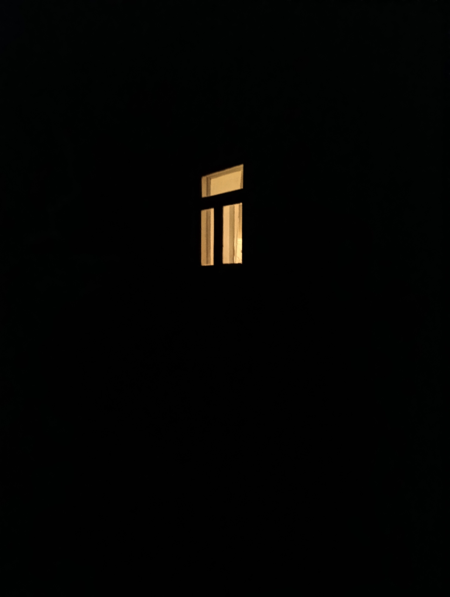 The image depicts a small, rectangular window with vertical panes, illuminated by a warm, yellowish light from within. The window is set against a backdrop of deep blackness, suggesting it is night or an interior space with minimal ambient light. The window is positioned centrally in the upper half of the frame. The light emanating from the window creates a stark contrast with the surrounding darkness, drawing the viewer's attention to this single point of illumination. There is no information in the image to suggest a location in Roghi, Moldova, or any details about people, actions, interactions, or the environment beyond the window.
