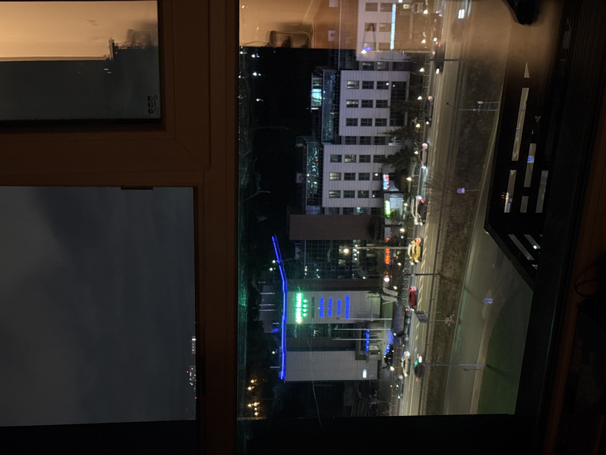 The image is a view from a window at night in Vidnoye, Russia. The window frame is light brown wood. Outside, a street with moderate traffic can be seen with buildings.
A tall, white building with multiple rows of windows is visible. Another building displays blue and green neon lighting. Streetlights illuminate the road. The sky is dark, indicating nighttime. The window reflects some light from the interior.