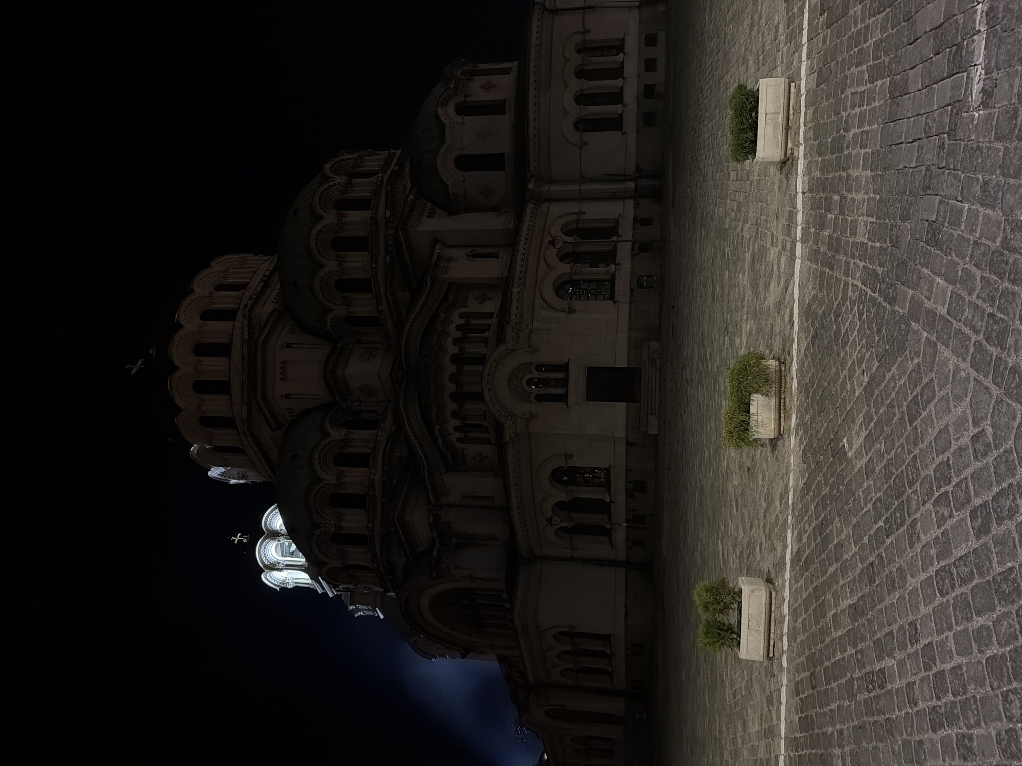 The image shows the exterior of St. Alexander Nevsky Cathedral in Sofia, Bulgaria, at night. The cathedral is a large, domed Orthodox church constructed primarily of stone and is illuminated by artificial lights, casting a dramatic shadow across the cobblestone plaza in front of it. The sky is dark and appears clear. Three large, rectangular planters containing greenery are positioned on the plaza, spaced apart and facing the cathedral. The cobblestone pavement is laid in a uniform pattern. There are no people or vehicles visible in the frame. A beam of light, possibly from a spotlight, is visible on the left side of the building, illuminating a portion of the facade and creating a hazy effect. The overall scene conveys a sense of solemnity and grandeur.