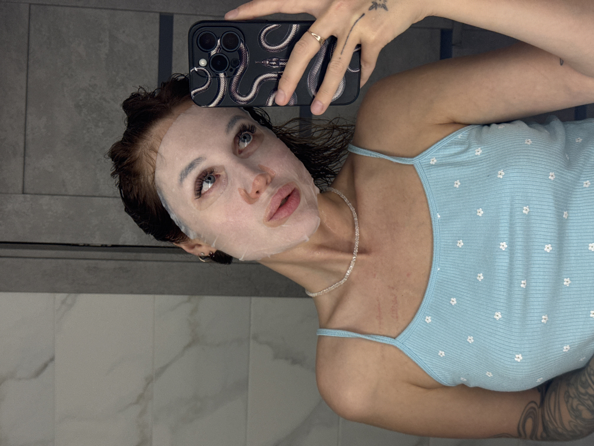 A young woman with dark red hair, wearing a light blue ribbed tank top with a floral pattern and a delicate pearl necklace, is taking a selfie in a bathroom. Her face is covered with a white sheet mask, and she is holding up a smartphone with a black snake-patterned case. The background shows tiled walls. The image does not provide any information about the location in Chaikovsky, Russia.