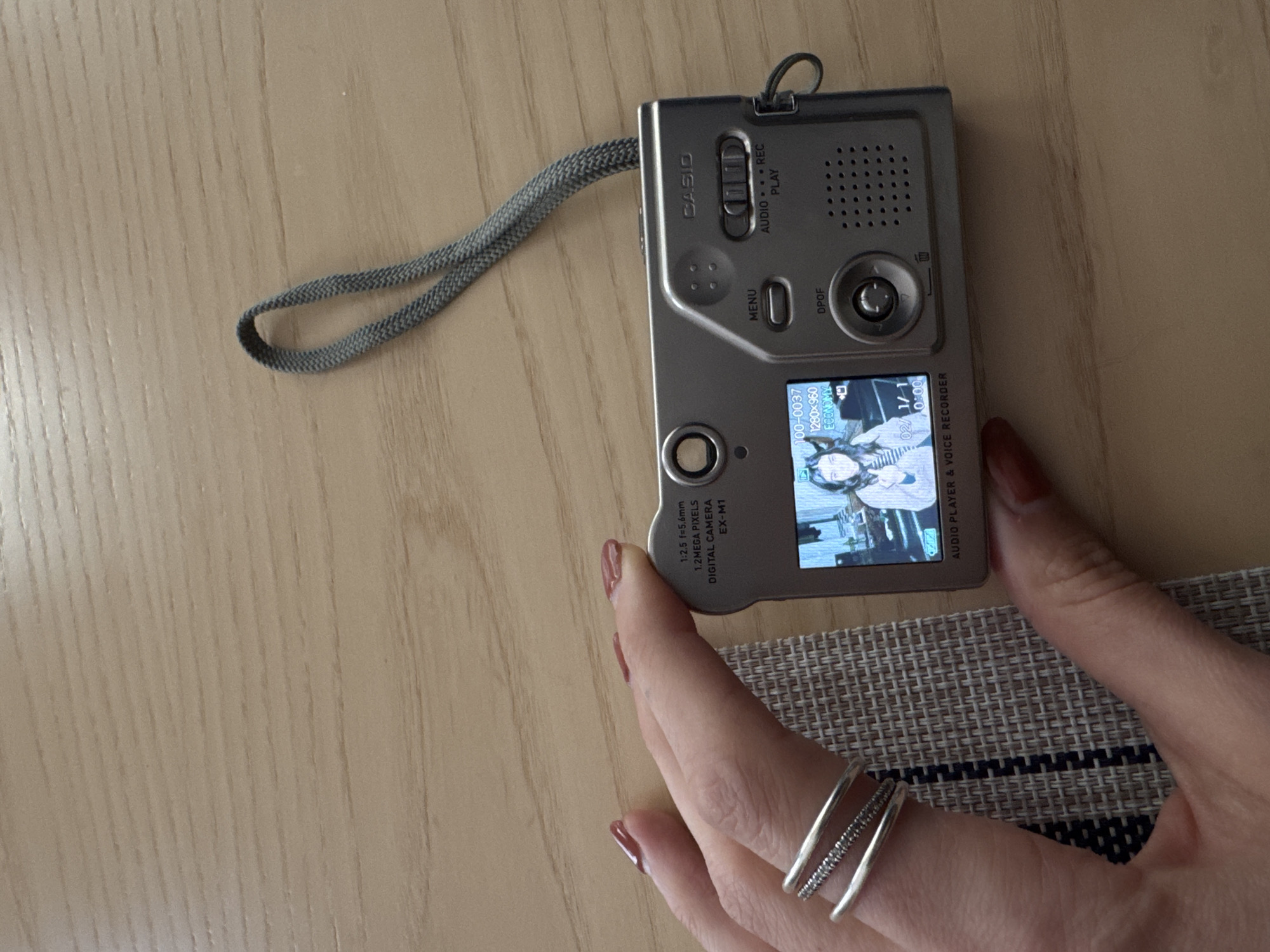 A close-up shot shows a Casio EX-M1 digital camera resting on a surface, possibly a table, with a hand partially holding it. The camera's screen displays a video or image, with identifiable settings and information visible on the screen and body of the camera. A gray strap is attached to the top of the camera, extending towards the left side of the image. The hand features multiple silver rings. A textured mat or surface is visible near the hand, providing a backdrop against the light-colored wooden surface. There are no obvious indicators of the location being in New York City.