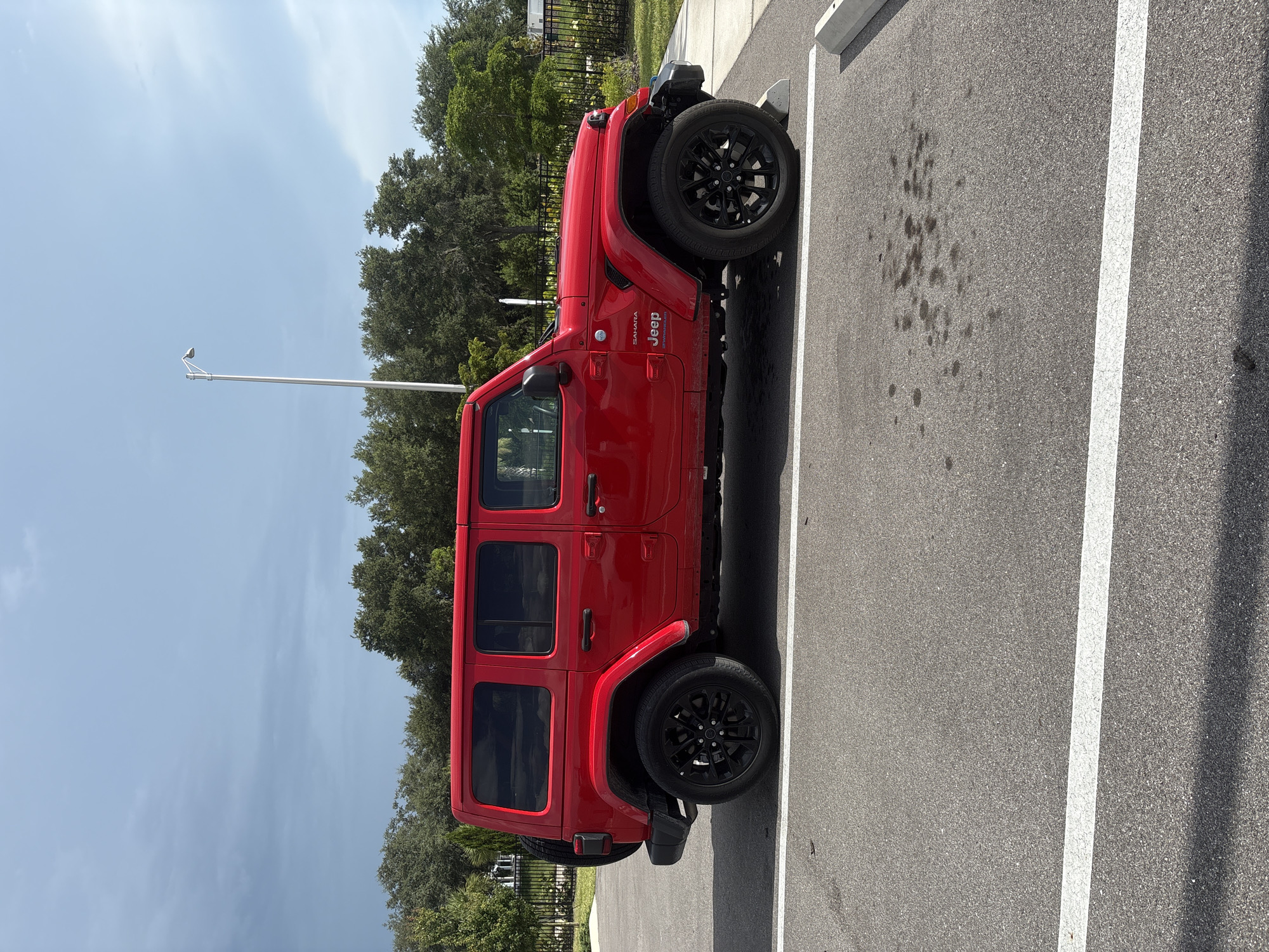 A red Jeep Wrangler Sahara is parked in a lot with a light grey sky. The vehicle is parked in a marked parking space with white lines. The Jeep is a four-door model with black rims. Behind the Jeep, there is a row of trees and a black metal fence. In the distance, there are buildings and a street lamp. The ground is asphalt with a few dark stains. The image appears to be taken in Sarasota, Florida, United States.
