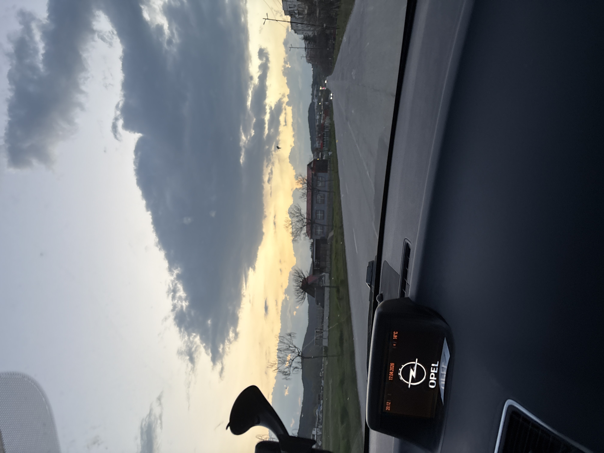The image, taken from inside a car, shows a view towards the city of Panagyurishte, Bulgaria. The car's dashboard is visible, including a display indicating the Opel car brand and a temperature of 14°C. The sky is overcast with clouds, and the setting sun casts a golden glow near the horizon. A road stretches into the distance, lined with buildings and trees. In the background, a mountain range is visible. A phone mount is seen affixed to the car's dashboard.