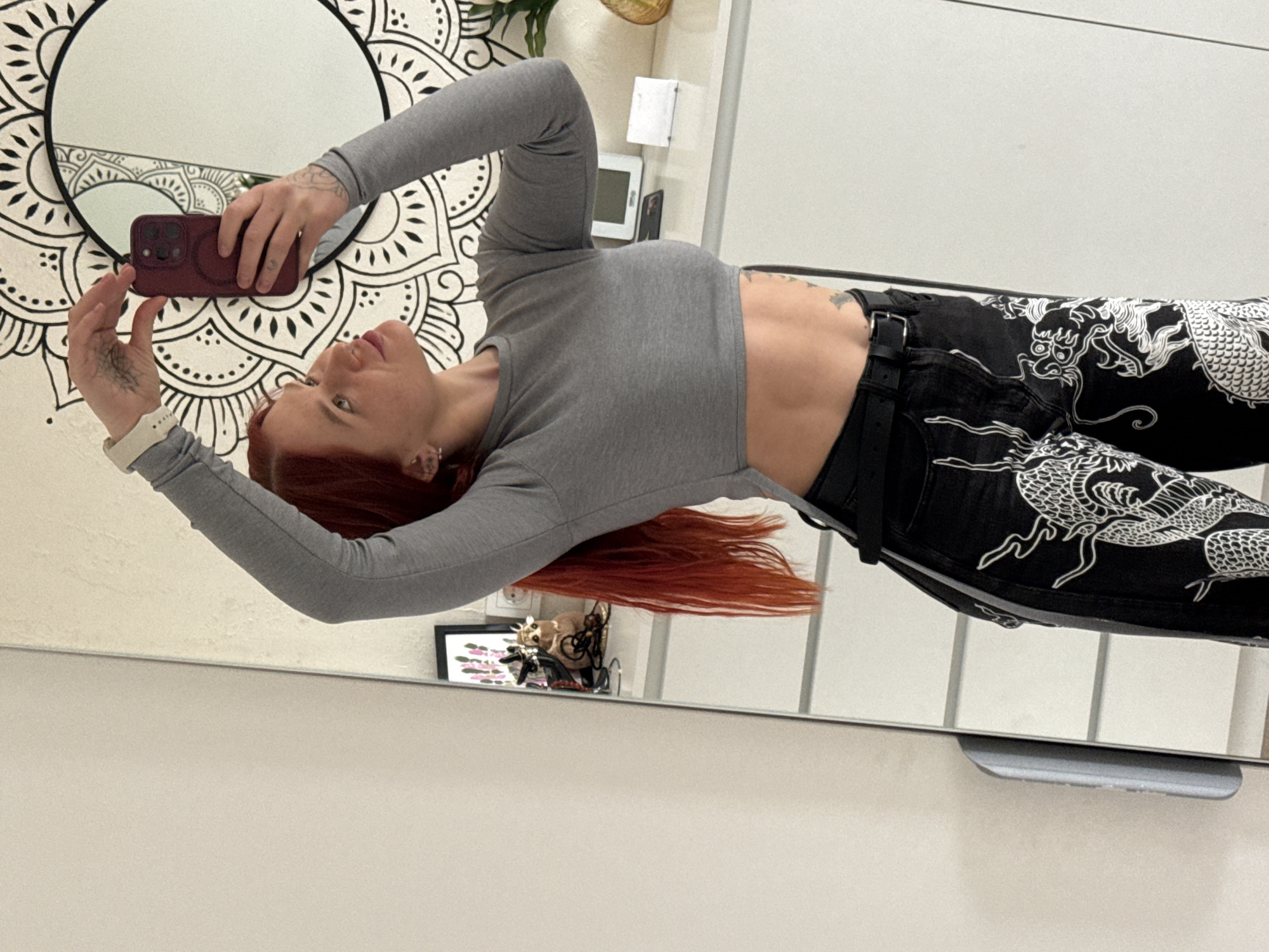 A woman with red hair, wearing a gray long-sleeved crop top and black pants with a white dragon pattern, takes a selfie in a mirror. The mirror is circular with a mandala design on the wall behind it. The mirror is reflecting the room, which includes a white cabinet, framed artwork, and decorative items. The photo appears to have been taken indoors. The location in the city of Rostov-on-Don, Russia is not verifiable from the image.