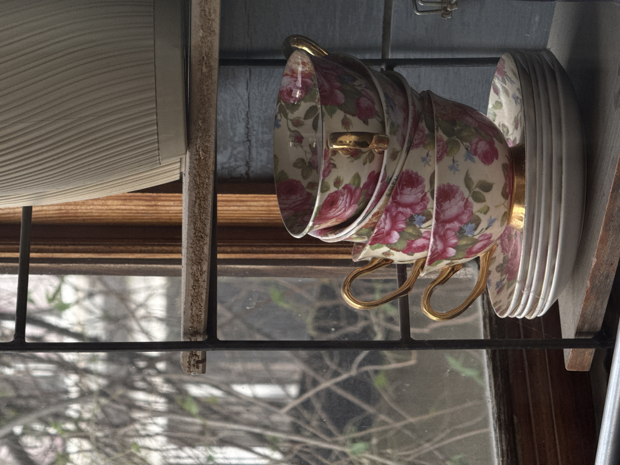 The image shows a stack of four vintage teacups with a floral pattern, featuring pink roses and blue accents, on a wooden shelf. The cups have gold handles and rims. Below the cups, a stack of saucers with the same floral pattern is visible, topped by a white saucer. Behind the teacups and saucers, a window with bare branches of trees is visible, suggesting an outdoor scene. The shelf is part of a multi-tiered shelving unit, with another shelf above it holding a ribbed, beige planter. The overall scene evokes a rustic, vintage aesthetic. The location is not specified in the image.