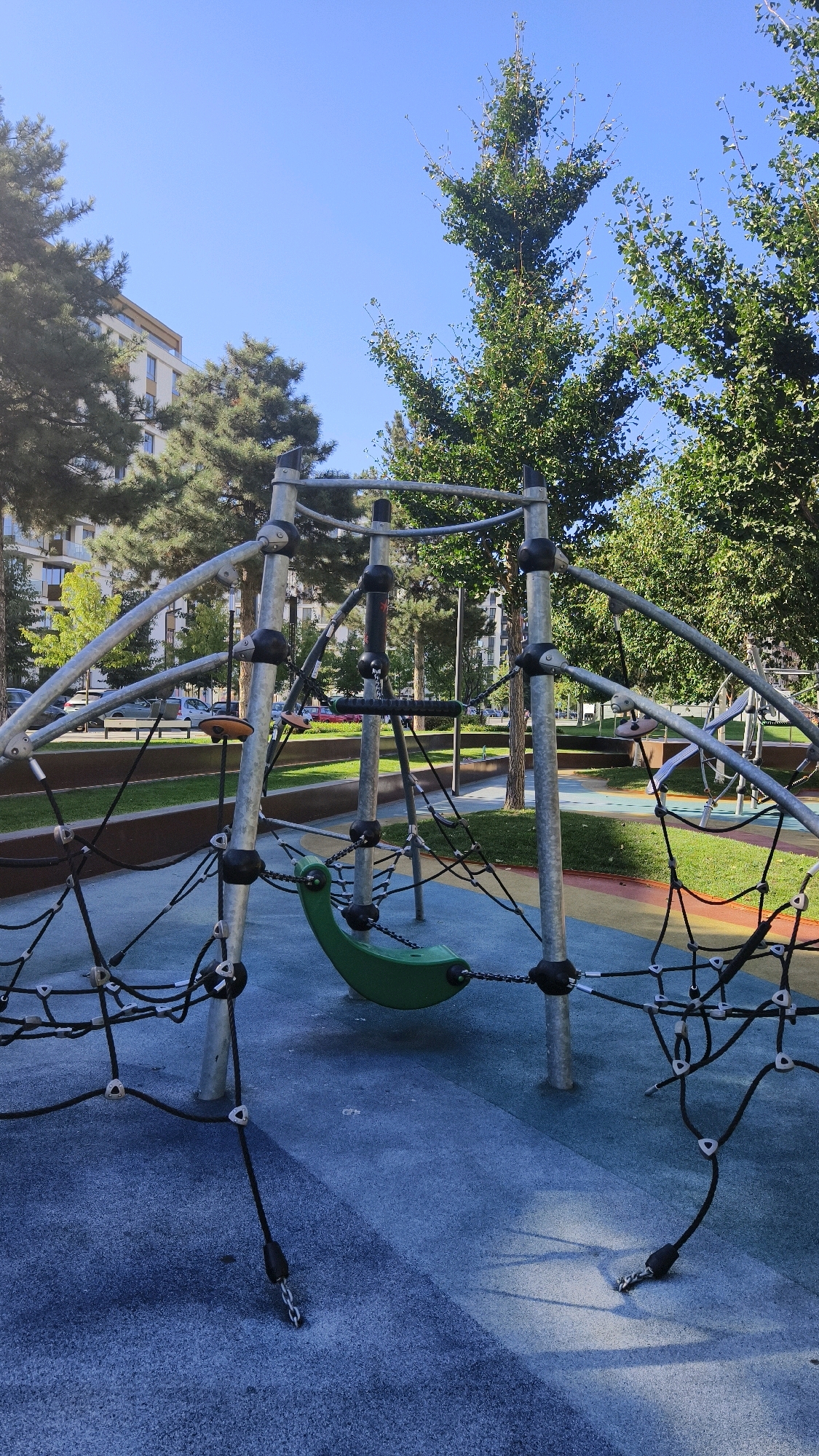 This is a vertical, eye-level, outdoor, daytime photograph of a modern children's playground. The playground features a large, metallic climbing structure with a green, suspended seat. The ground is a soft, rubberized surface with blue and grey tones, marked with yellow and red lines in the background. Surrounding the playground are several trees with lush green foliage and a clear blue sky overhead. In the background, a multi-story building with a light-colored facade is visible, along with parked cars on a street. The scene is set in the city of Belgrade, Serbia, specifically in the New Belgrade area, known for its contemporary architecture and green spaces. There are no people visible in the frame, and the playground appears to be unoccupied. The sunlight casts distinct shadows on the ground, indicating a bright, sunny day. The overall impression is one of a well-maintained, urban recreational area.