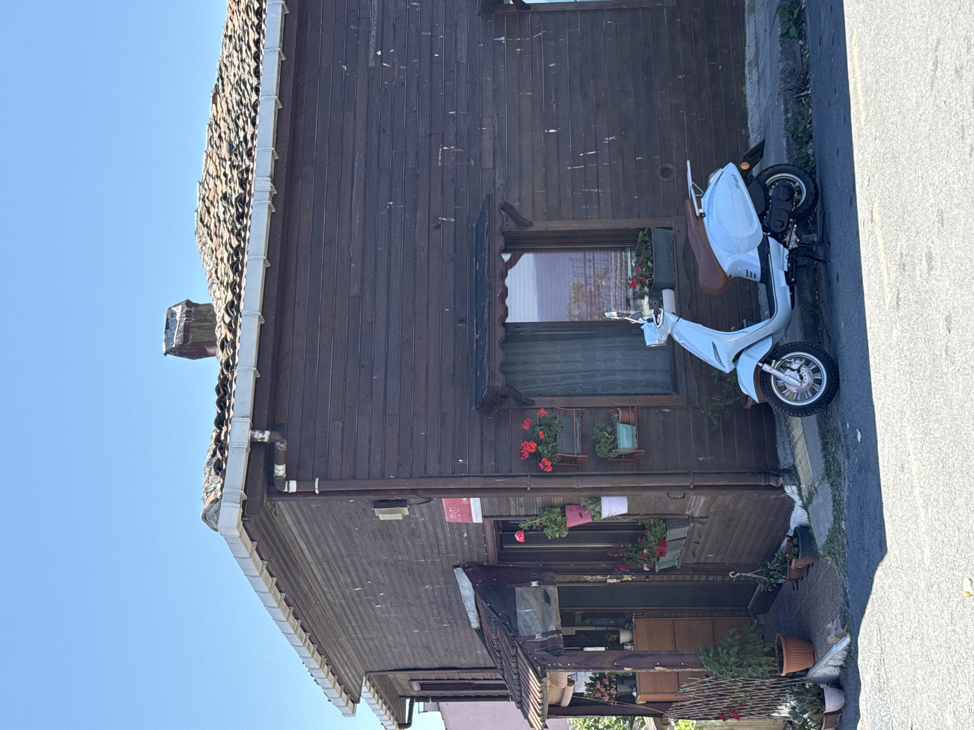 A light blue scooter is parked on the side of an asphalt road in front of a two-story wooden building. The building has a dark brown wooden exterior with horizontal planks and a tiled roof. There are several windows on the facade, some adorned with flower boxes containing red and pink flowers. A small awning covers the entrance to the building, which is flanked by potted plants. A street sign is visible on the side of the building. The sky above is clear and blue. The scene appears to be in a residential or commercial area.
