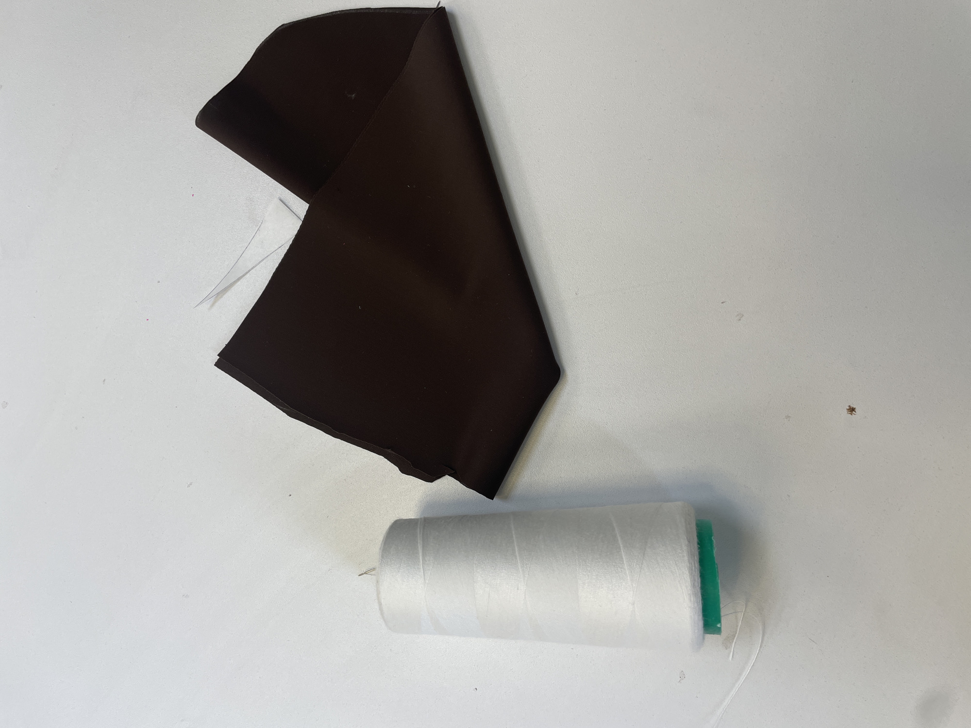 The image shows a spool of white thread with a green base, lying on a white surface. To the right of the spool, there is a folded piece of dark brown fabric. The fabric appears to be shiny and smooth. A single strand of white thread is visible extending from the spool and slightly onto the fabric. The background is a plain white surface, with some faint marks and specks visible. The lighting suggests an indoor setting. The image does not provide any information that would allow for its location in Sinyavino, Russia.