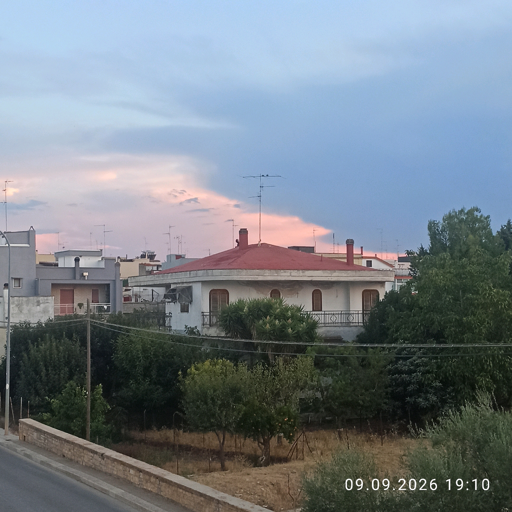 This is a photograph of a residential area in Bitonto, Italy, taken at dusk. The sky is a gradient of pale blue transitioning to a darker blue and pinkish clouds. Several buildings are visible, including a prominent white house with a red roof and a balcony. Various antennas are mounted on the rooftops. The foreground features a low brick wall bordering a street or pathway. Trees and vegetation are scattered throughout the scene, with some dry grass and a few small orange fruits on a tree. The time stamp on the image indicates it was taken on September 9, 2026, at 19:10. There are no people visible in the image.