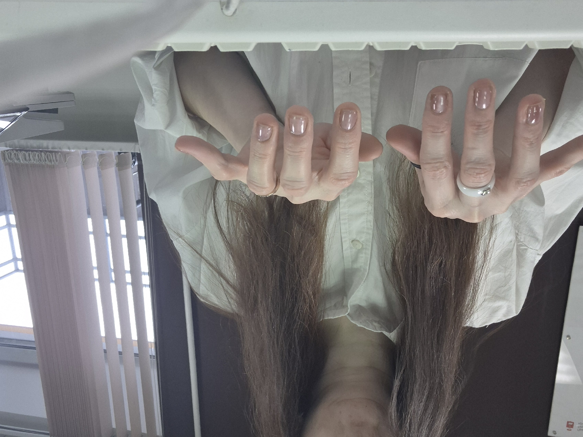 The image depicts a woman from a close, inverted perspective, showcasing her hands and upper torso. She is wearing a white button-down shirt and has long, brown hair. Her hands are adorned with minimalist clear nail polish and two rings - a black band on one hand and a white ring with a stone on the other. The background reveals a room with blinds and a partially visible window to the left, and a white surface with unknown markings or text to the right. The location is specified as Unknown, Unknown.