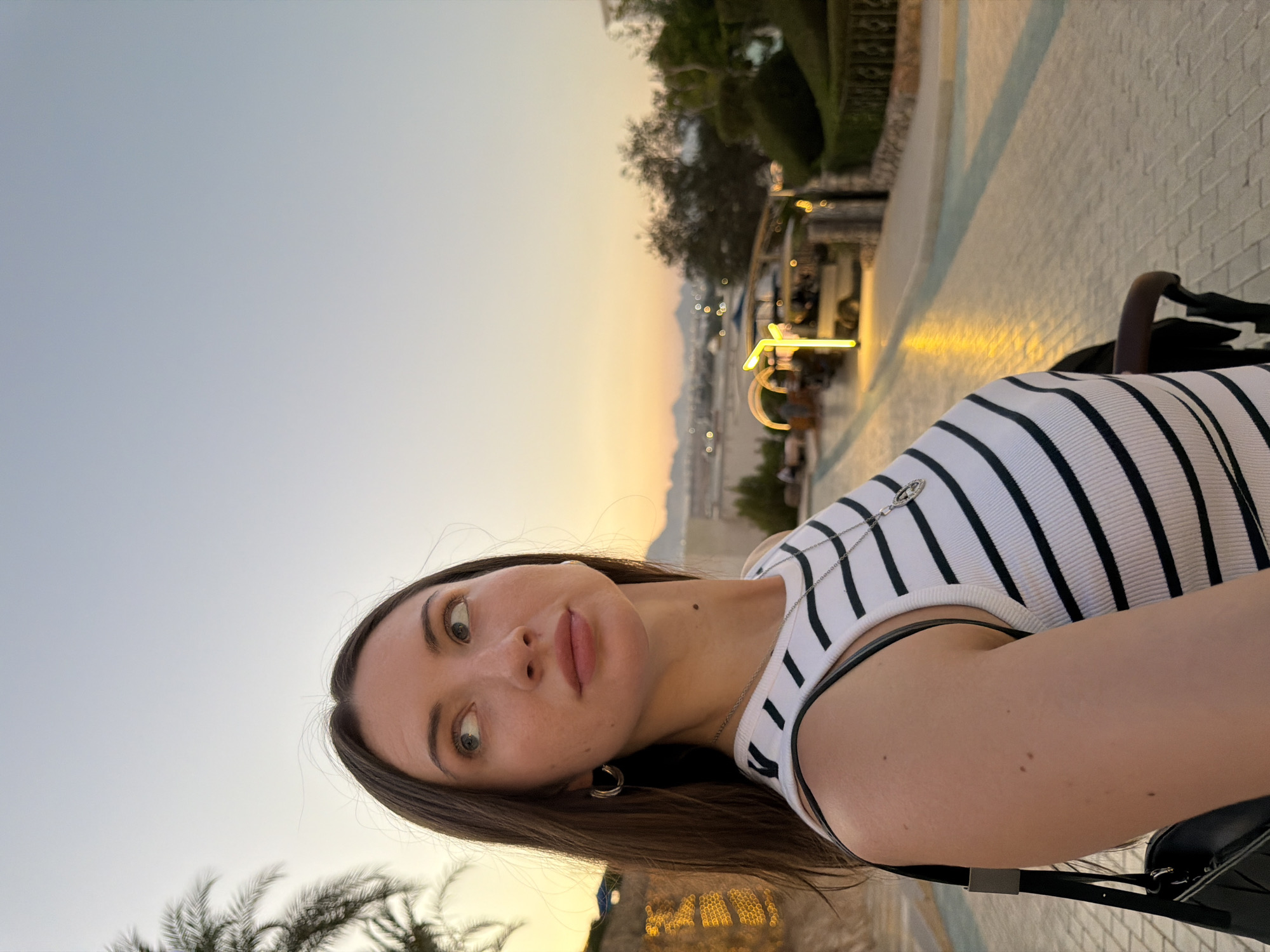 A young woman with long, dark brown hair is pictured in a selfie-style shot. She is looking upwards and to the right, away from the camera. She is wearing a black and white striped tank top and a silver necklace with a pendant. A silver hoop earring is visible in her left ear. She has a mole on her neck.
The background suggests an outdoor setting at dusk, possibly in Sharm El Sheikh, Egypt. Palm trees are visible on the left. A paved walkway with soft yellow lighting lines it. In the middle ground, there are several small vehicles, possibly golf carts or service vehicles, and some landscaping. Further in the background, there are distant hills or mountains, hinting at a coastal or desert landscape. The sky is a pale gradient of orange and blue, indicative of sunset or twilight. The overall impression is a casual, outdoor evening scene in a resort or tourist area.