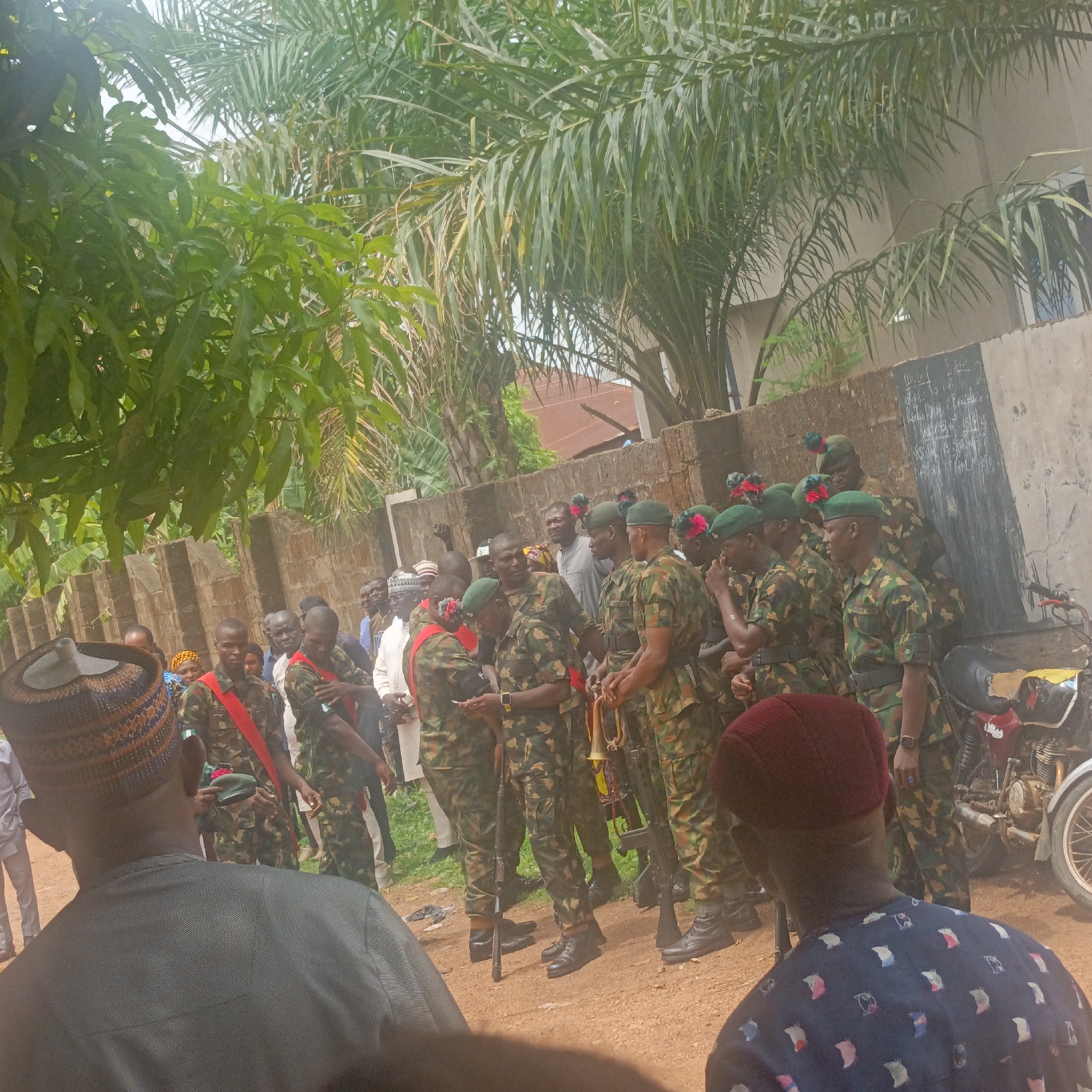 The image depicts a group of soldiers in camouflage uniforms standing on a dirt ground in Akwanga, Nigeria. The soldiers wear green berets with decorations, red sashes, and black boots. They hold rifles and trumpets. Behind the soldiers, there's a gathering of civilians. A motorcycle is parked on the side. The backdrop includes trees and a wall.