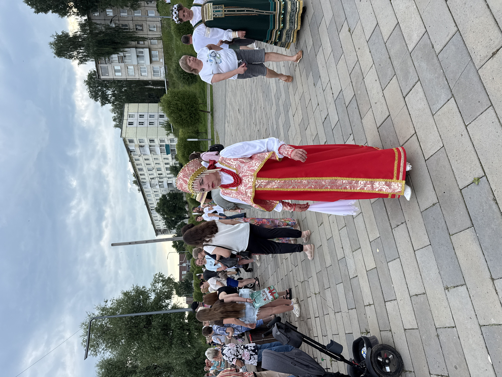In Votkinsk, Russia, an outdoor scene unfolds on a paved area near buildings and trees. A woman in a traditional red and gold costume, complete with an elaborate headdress and beaded necklace, stands prominently in the foreground. She is looking to her left. To her right, another woman in a green dress with gold trim and a floral crown is also present. Behind them, a crowd of people are gathered, some walking and others standing, engaged in various activities. The environment includes trees, streetlights, and residential buildings. A stroller is visible on the left side of the image. The sky is overcast with clouds.