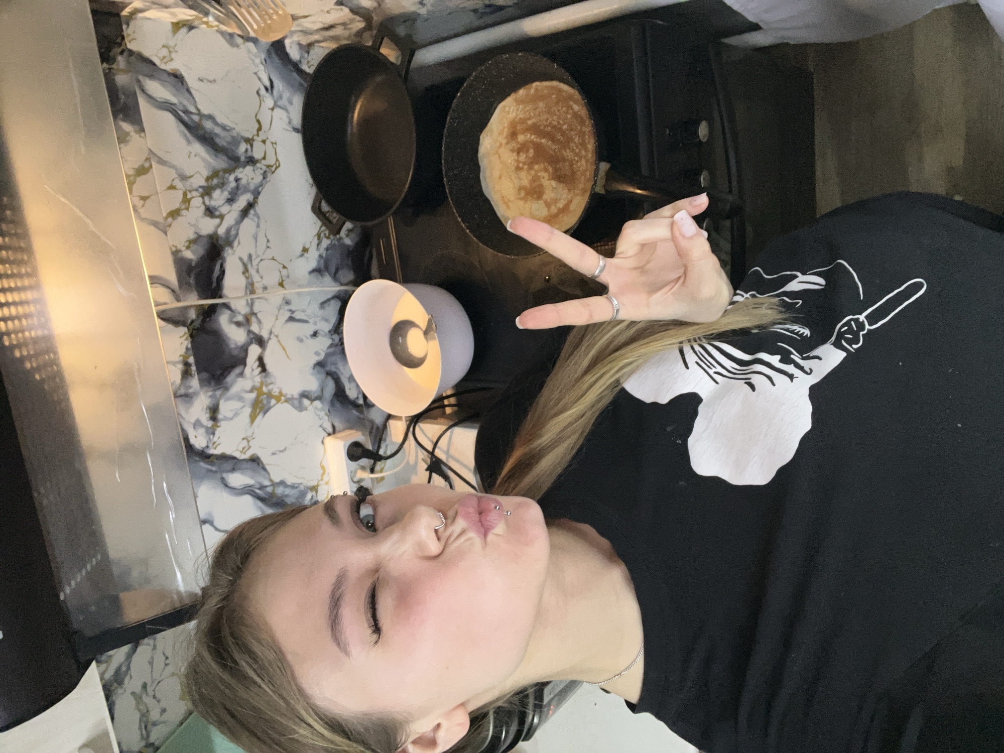 A woman is standing in a kitchen, facing the camera, and making a peace sign with her right hand. She is wearing a black t-shirt with a white design. Her face is turned to the left, with her eyes closed and lips pursed. She has multiple piercings on her face, including her nose and eyebrow. Her hair is blond. On the stove behind her, there is a frying pan with a pancake cooking. Next to it is another empty frying pan. There is a lamp next to the stove. The kitchen has a white and gray marble countertop and is located in the city of Фоки, Russia.