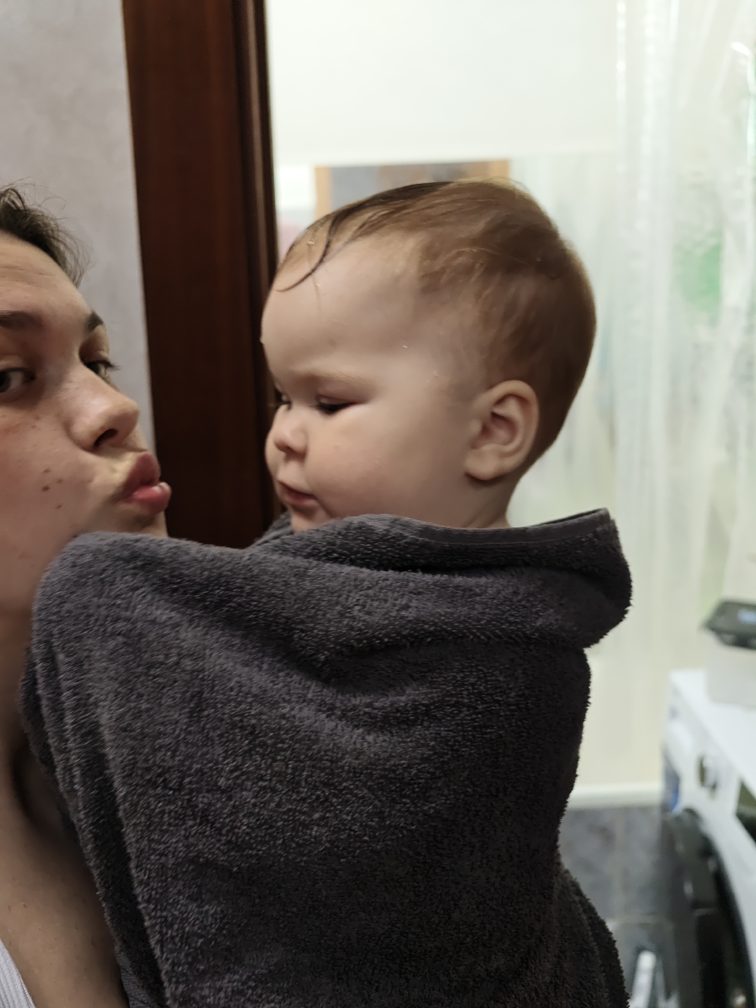 The image shows a person holding a baby wrapped in a gray towel, likely after a bath. The person has their lips puckered as if giving a kiss to the baby, who is looking off to the side. The background shows what appears to be a bathroom setting, with a washing machine visible on the right. A dark wooden door is seen partially behind the person and baby on the left. Based on the prompt, this scene is likely located in Chaikovsky, Russia.