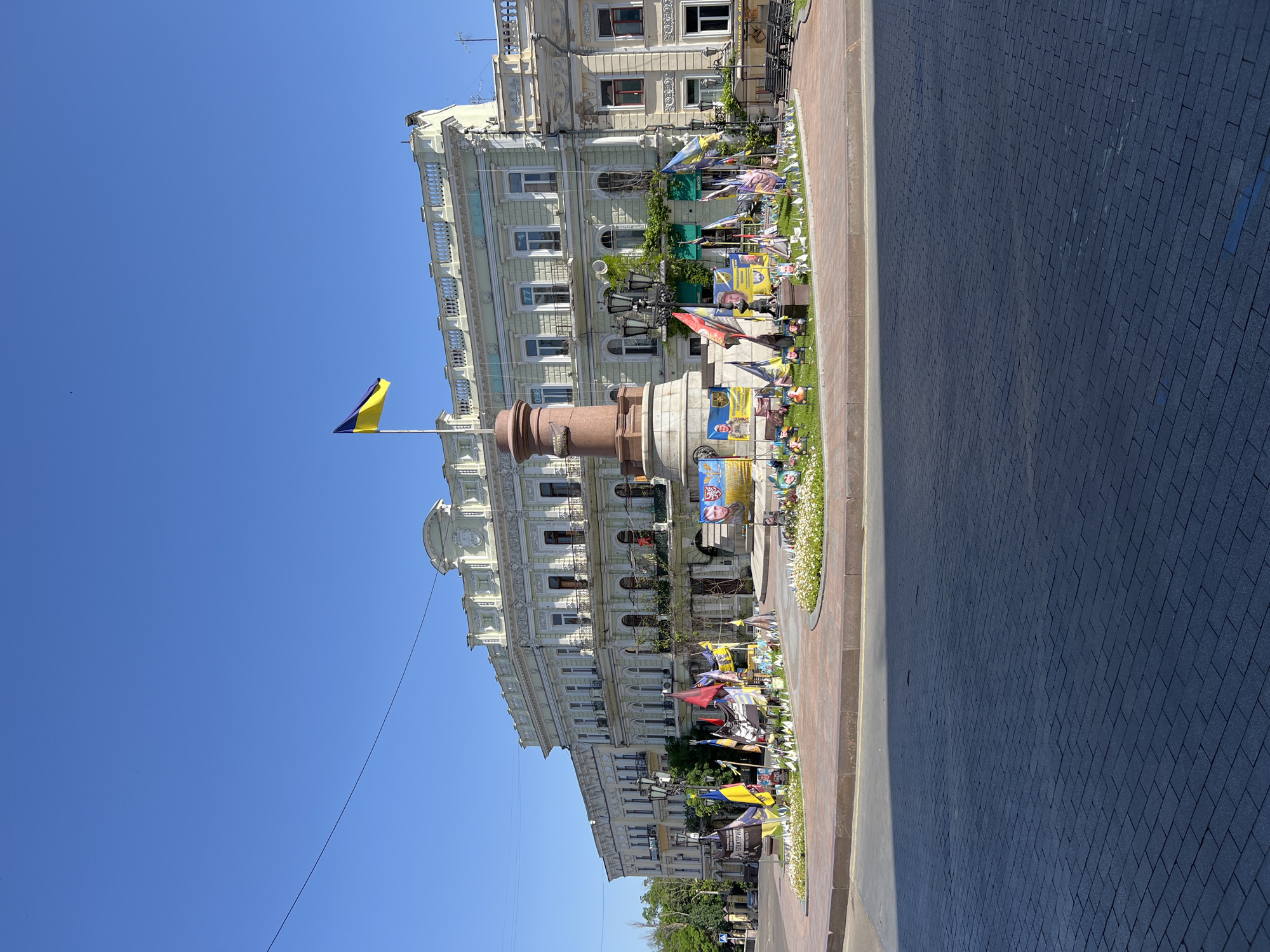 The image depicts a street scene in Odesa, Ukraine, featuring a monument topped with a Ukrainian flag in front of a large, ornate building. The monument is surrounded by smaller Ukrainian flags and what appear to be posters or banners. The building behind the monument is multi-storied with decorative architectural details, including balconies and window frames. The street is paved with dark-colored blocks. Clear blue sky is visible above the building. The image is taken at street level, and the orientation is rotated 90 degrees.