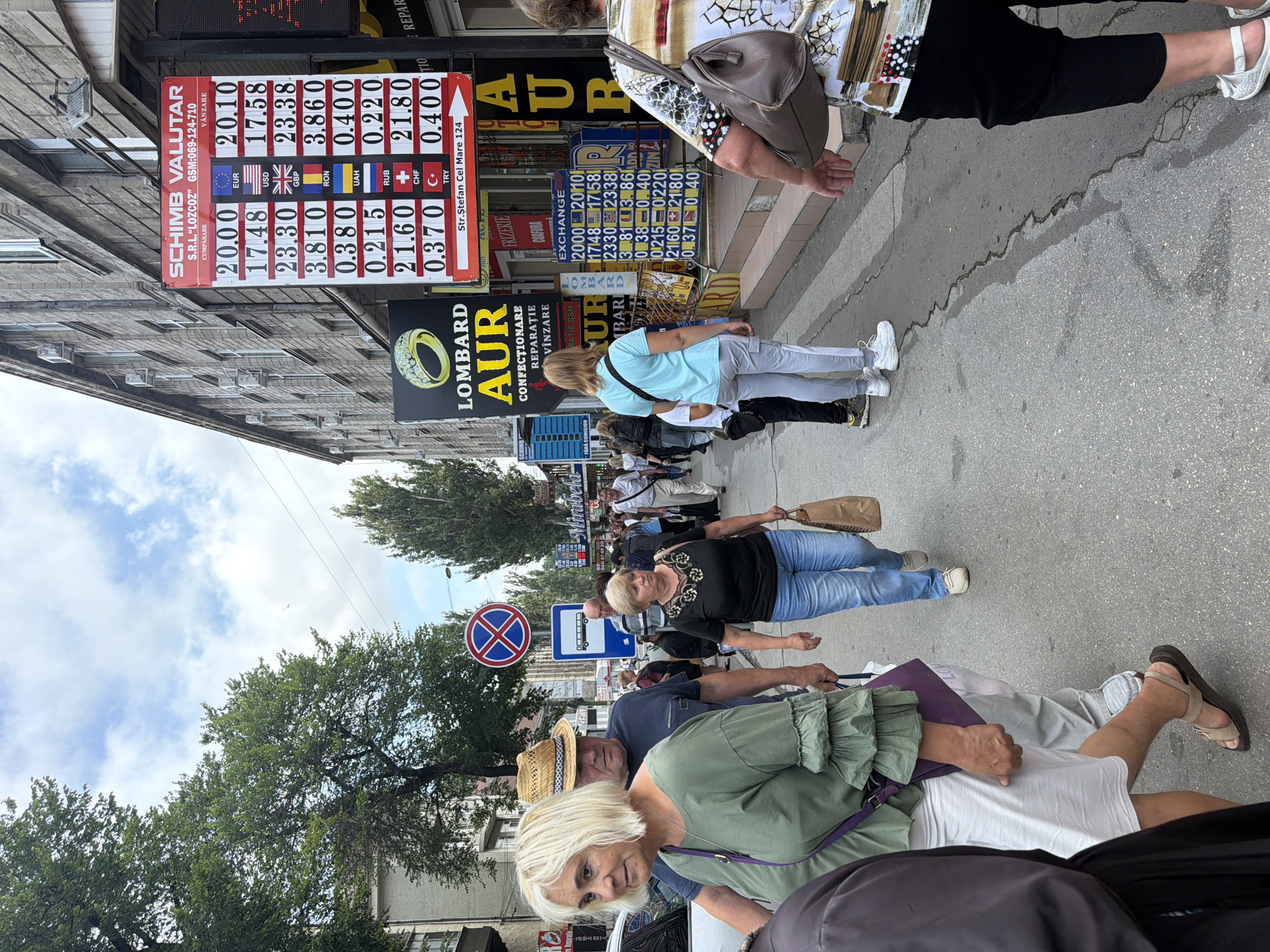 This image shows a street scene in Chișinău, Moldova, specifically on Ștefan cel Mare Boulevard, as indicated by a sign visible on the right side of the frame. A currency exchange office, labeled "SCHIMB VALUTAR," is prominently featured with a large exchange rate board displaying prices for EUR, USD, GBP, RON, UAH, RUB, CHF, and TRY. Next to it is another exchange board with similar information. Several people are walking along the sidewalk, some looking at the exchange rates. A woman in a green top and white skirt is in the foreground on the left, and a man in a blue shirt and straw hat is beside her. Further down the street, other individuals are visible, including a woman in a black top and blue jeans holding a paper bag. Traffic signs, including a "no parking" sign, are also present. The buildings lining the street are typical of urban architecture. The sky is partly cloudy.