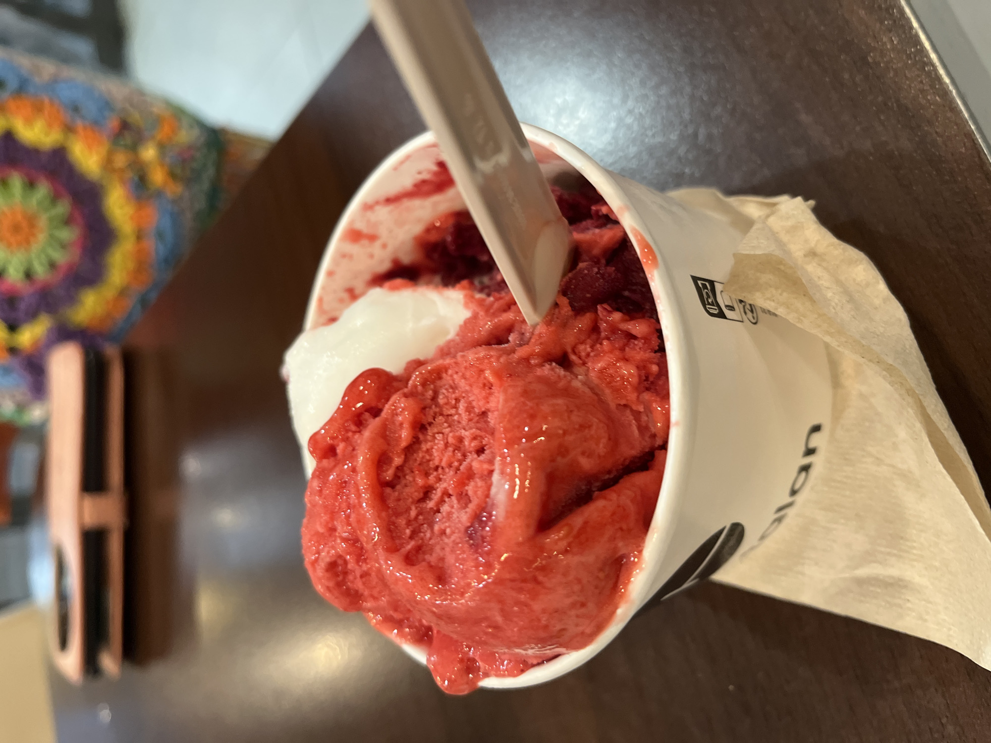 In Palavas-les-Flots, France, an ice cream cup sits on a dark wooden table, partially obscured by a crumpled light brown napkin. The cup, white with black text and a logo, contains two scoops of vibrant red sorbet and a dollop of white ice cream. A wooden spoon with white remnants of ice cream rests inside the cup, angled towards the foreground. In the background, a pink phone case is visible on the table, and a colorful crocheted item, likely a bag or blanket, is draped over something out of frame. The lighting suggests an indoor setting, possibly a cafe or shop.