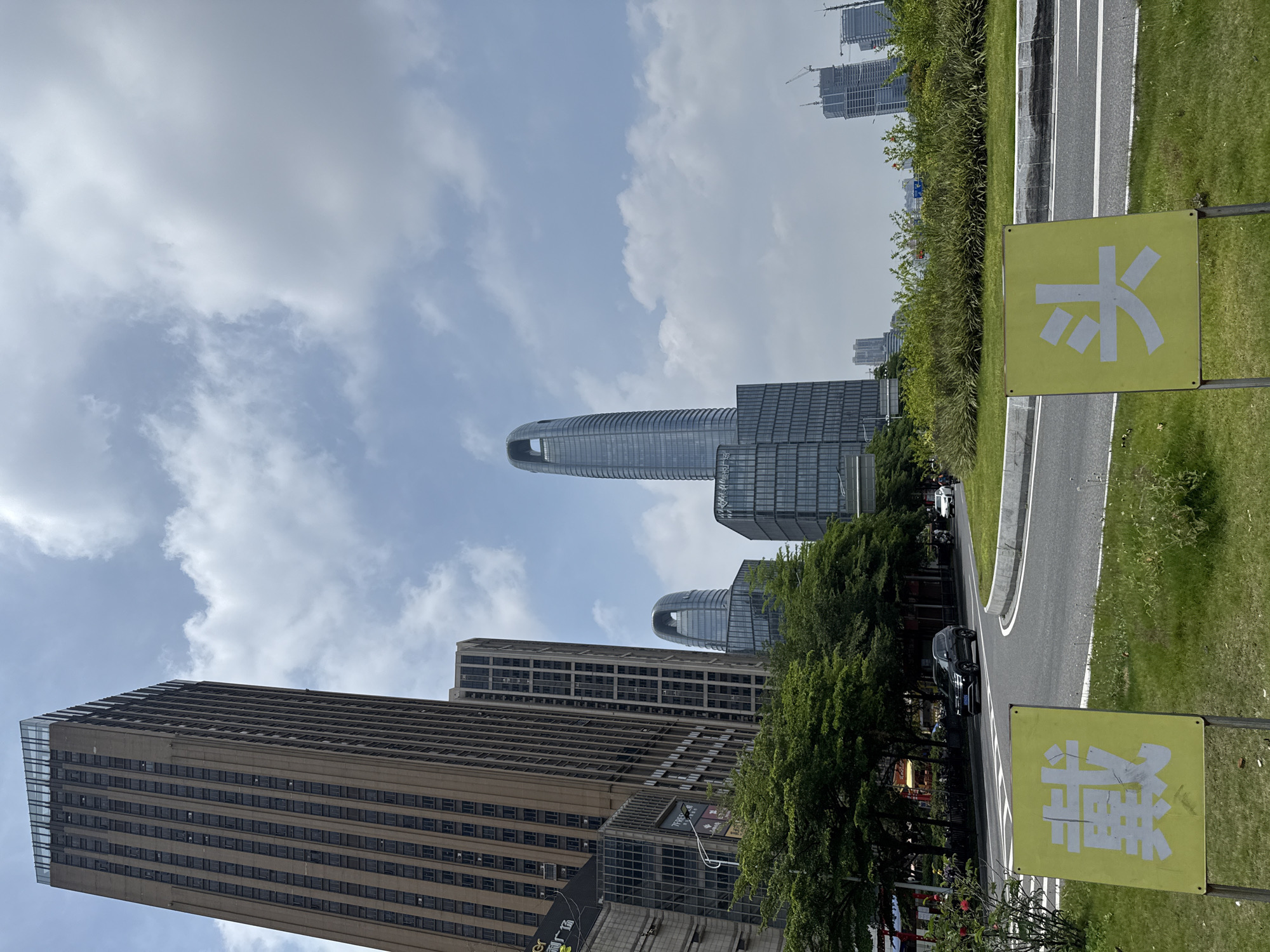 This image presents a detailed urban landscape in the city of Haizhu District, China, under a partially cloudy sky.
In the foreground, a grassy median separates a curved asphalt road. Within this median, two rectangular yellow signs with white Chinese characters are prominently displayed. The left sign shows "戴" (dài), and the right sign shows "头" (tóu). The grass is interspersed with small green plants. On the road, a black SUV is visible driving from left to right, and a white vehicle along with a motorcycle are partially seen further down the road on the right.
The mid-ground is dominated by architectural structures and dense foliage. A line of mature trees with lush green leaves borders the left side of the road. Behind these trees and to the left, a tall, slender skyscraper with a brown facade and numerous small, vertically aligned windows rises sharply, topped with a modern glass enclosure. Adjacent and slightly behind it, another tall building with a darker, more contemporary design is visible. Towards the center-right and mid-ground, two distinctive modern skyscrapers made of glass and steel stand out; both feature a large, oval-shaped aperture near their apex, with the taller one positioned further back.
The background reveals more urban development. On the far right, several buildings are under construction, indicated by visible cranes and exposed structural elements. The sky above is a light blue, interspersed with white and grey cumulus clouds. No specific human figures are clearly discernible in detail, though indistinct shapes consistent with people are present on sidewalks near the buildings.