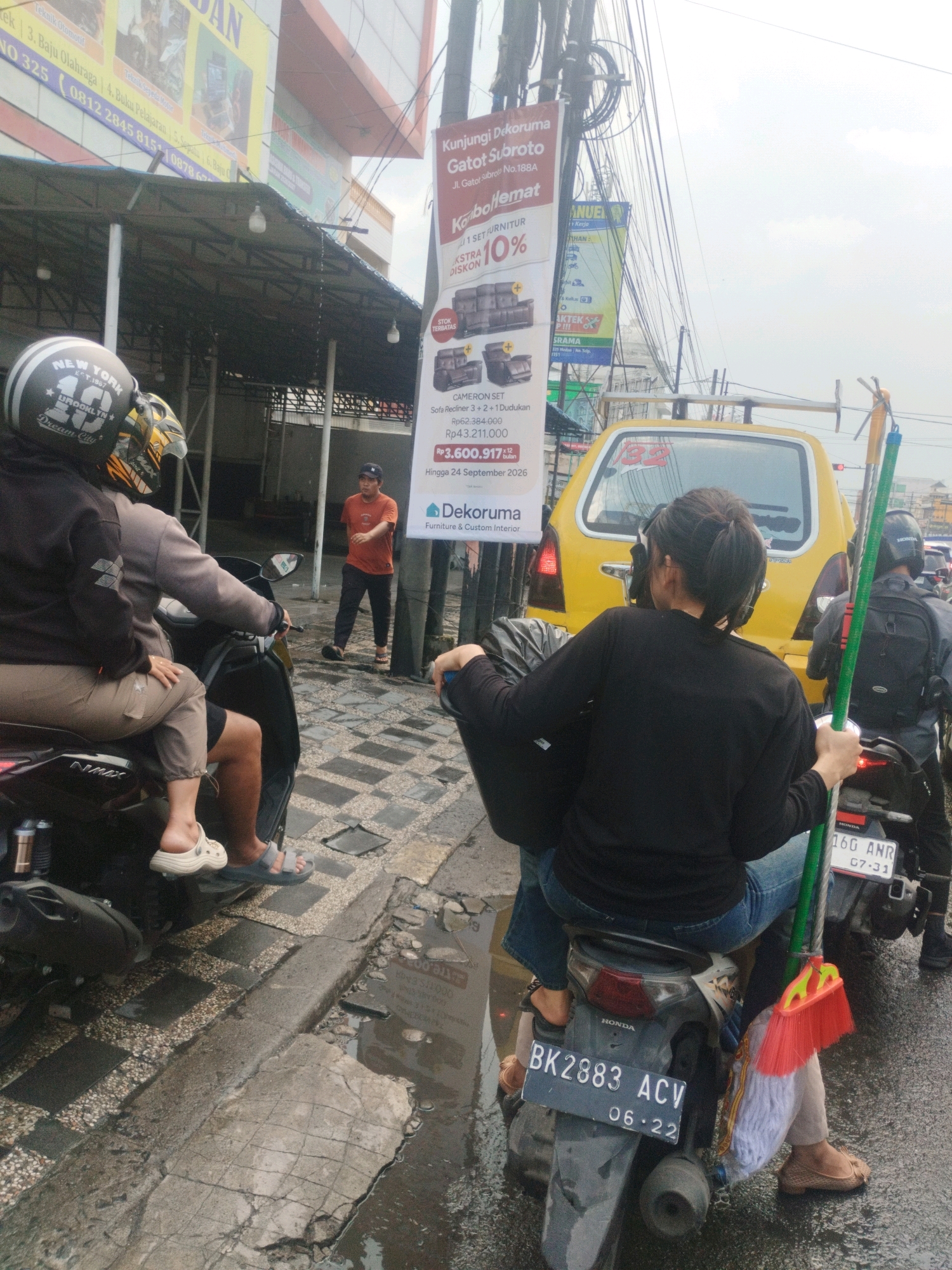 The image captures a street scene in the city of Medan, Indonesia, under an overcast sky. In the foreground, two individuals are riding on a motorcycle. The rider in front wears a black jacket and jeans, while the person behind them wears a black jacket and jeans, holding a black bag and a broom with a red brush. The motorcycle is a Honda, with license plate BK 2883 ACV, dated June 2022. To the left, another motorcycle is parked, with two people on it; the one in front wears a helmet with "NEW YORK" and "Dream City" text, and the person behind wears a helmet with a yellow and black design.
Further in the background, a yellow car with license plate 16G ANR is visible. A man in an orange shirt and black pants stands near a pole with a large banner advertising "Kunjugi Dekoruma Gatot Subroto" and a discount on furniture. The street appears wet, suggesting recent rain, with puddles and cracked pavement visible. Buildings, including what appears to be a shop with a large sign, line the background. Overhead, numerous electrical wires crisscross the sky. The overall atmosphere is that of a busy urban environment.