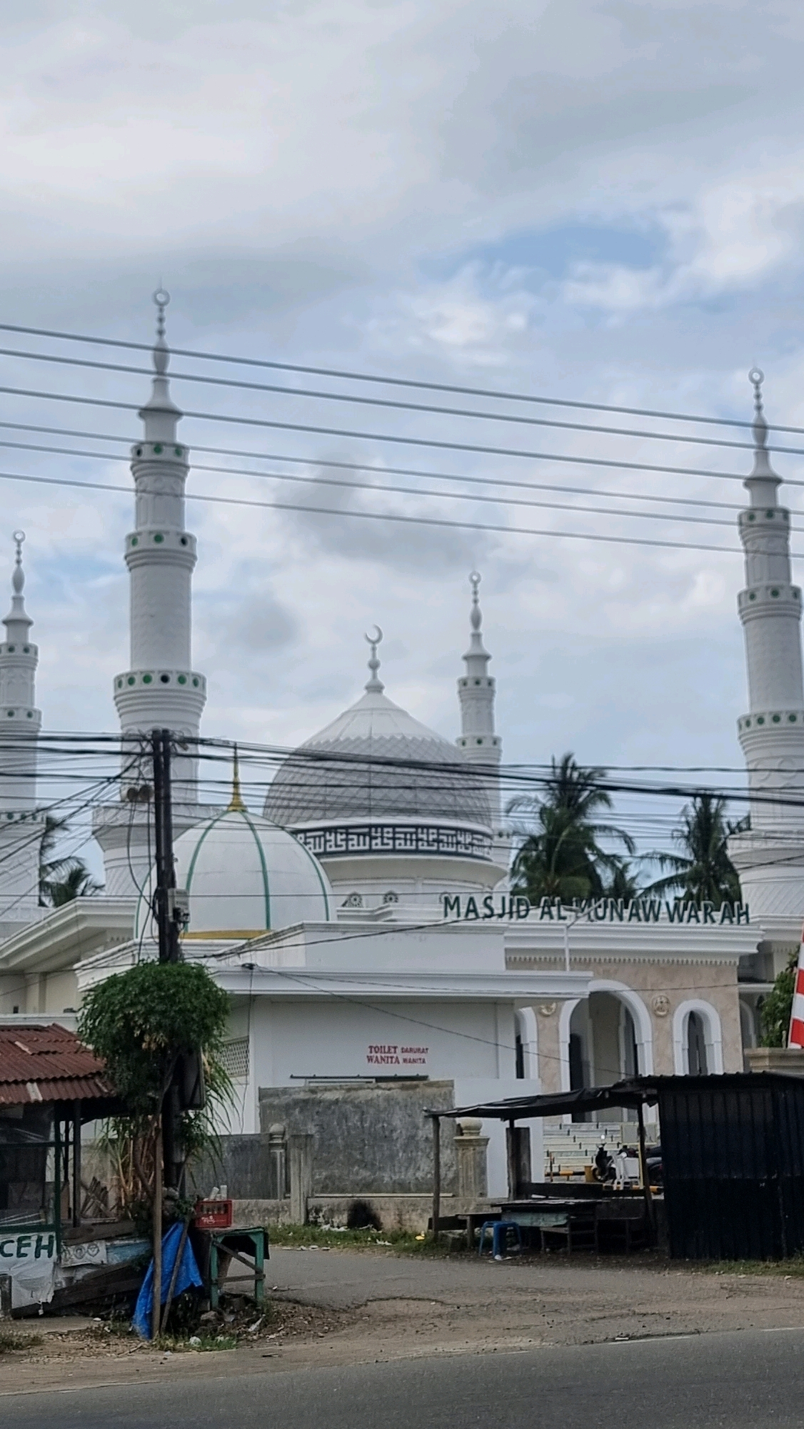 The image shows the Masjid Al Munawwarah in the city of Lhokseumawe, Indonesia. The mosque features white minarets and domes, with Arabic calligraphy on the main dome. In front of the mosque, there are several small roadside stalls and some utility poles with numerous wires crisscrossing the scene. A sign indicating "TOILET DARURAT WANITA WANITA" (Emergency Toilet for Women) is visible on a white wall. The sky is overcast with clouds.