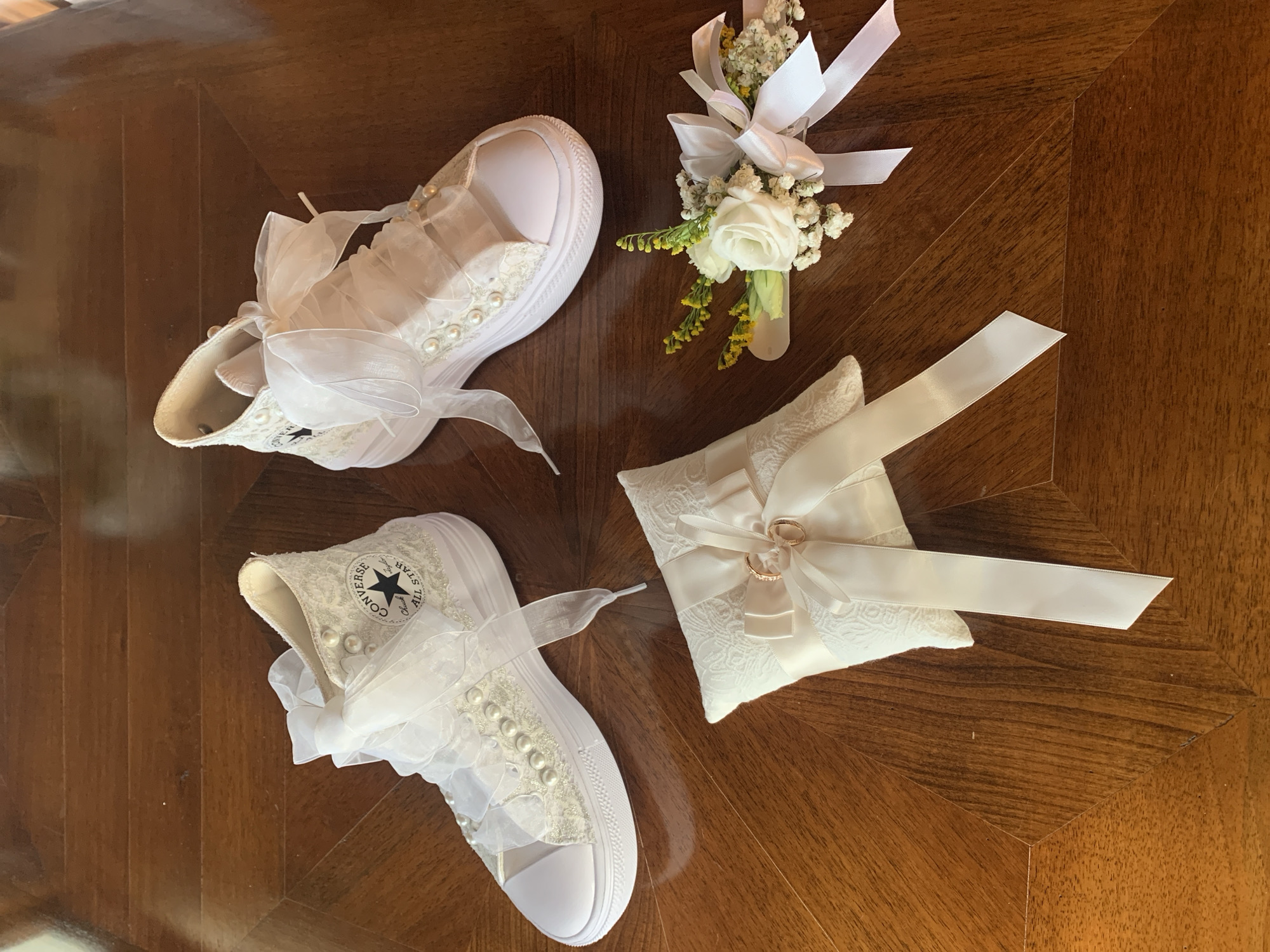 The image displays a collection of wedding accessories arranged on a polished wooden table. Two white Converse high-top sneakers, adorned with lace, pearls, and satin bows, are positioned at the top of the frame. In the lower left, a small white ring pillow with satin ribbons and two gold rings is visible. To the right of the pillow is a boutonniere featuring white roses, baby's breath, and yellow accents, tied with white ribbon. The overall impression is one of bridal preparation. The setting is not identifiable as Montepulciano, Italy, as no distinct landmarks or clues suggesting this location are present in the image.