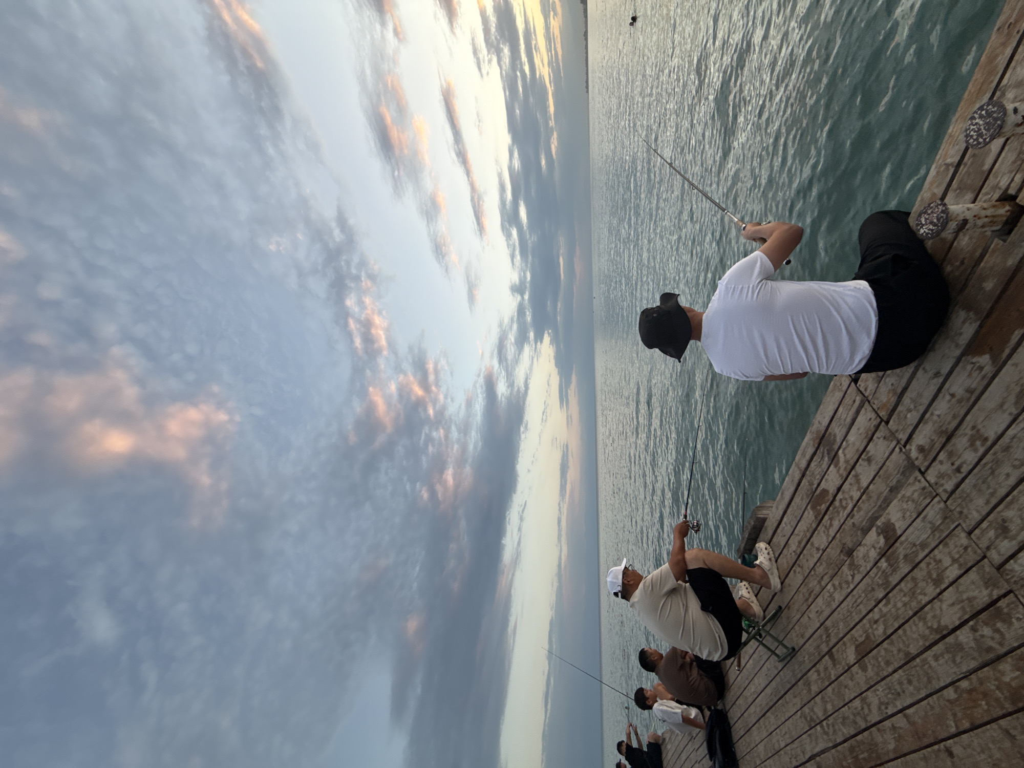 The image shows a group of people fishing from a wooden pier on a body of water, likely Issyk-Kul Lake, near Sary-Oy, Kyrgyzstan. The sky is filled with scattered clouds, tinged with pink and orange from the setting sun, casting a soft light on the scene. Several individuals are seated or squatting on the pier, holding fishing rods and casting lines into the water. One man in the foreground, wearing a white t-shirt and black shorts, is actively fishing, as is a man in a light-colored shirt and white cap to his left. Other individuals are also visible on the pier, engaged in the same activity. The water appears calm, with gentle ripples reflecting the sky. The wooden planks of the pier show signs of wear and tear, indicating its age and frequent use. The overall atmosphere is one of peaceful recreation as dusk settles over the landscape.