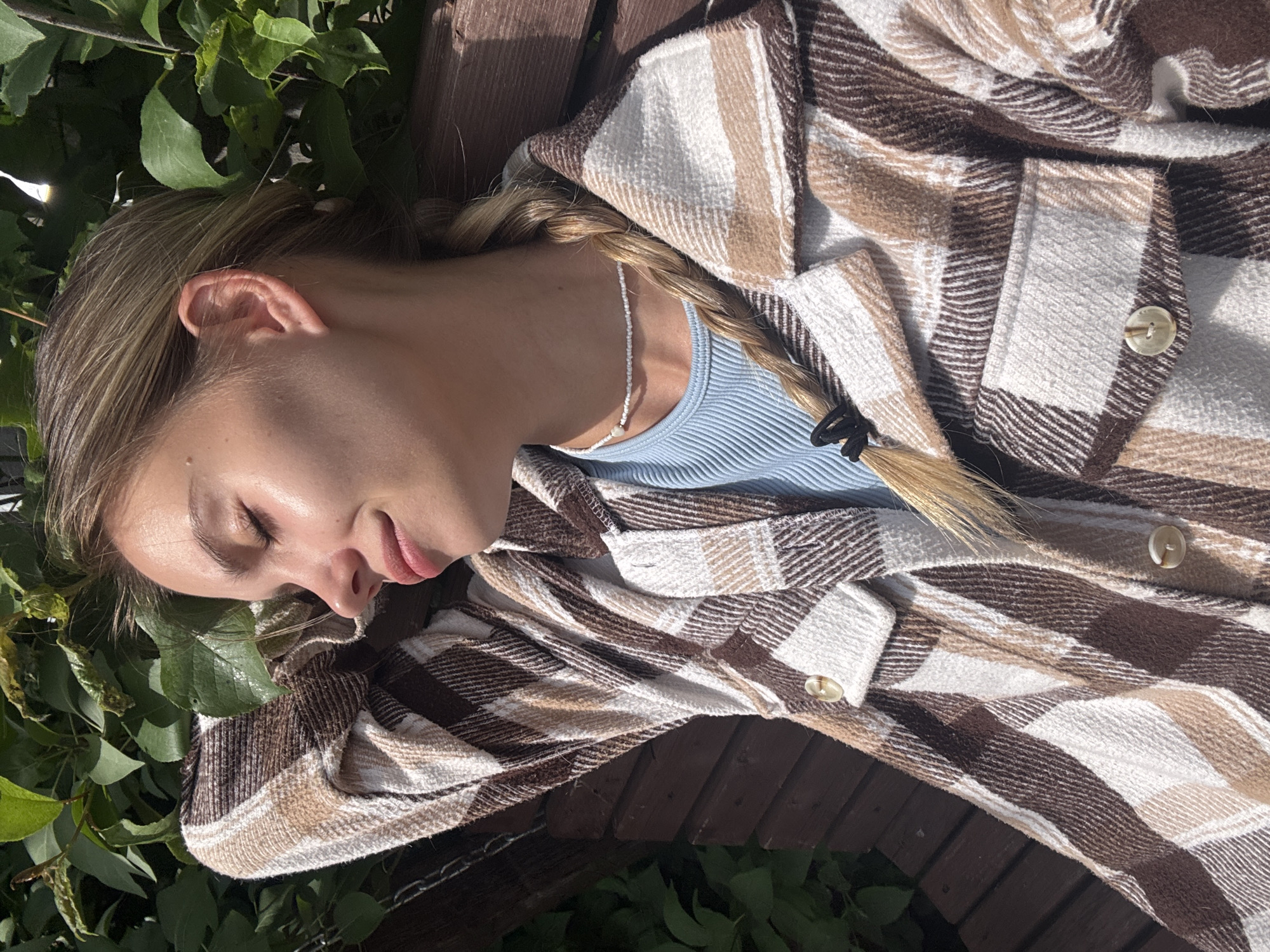 An outdoor, close-up shot captures a young woman resting with her eyes closed, conveying an impression of peace and relaxation. She is lying on her side, possibly on a wooden bench or railing, indicated by the visible dark brown wooden planks behind her. Her fair skin is illuminated by bright natural light, suggesting a clear, sunny day, likely late morning or afternoon, given the shadows and highlights.
The woman has light-colored hair, possibly blonde or light brown, styled into a single braid that extends over her shoulder and down her back. She is dressed in a light blue ribbed top, visible at her neckline, and a thick, long-sleeved plaid jacket or shirt featuring a pattern of brown, beige, and white squares. Several light-colored buttons are visible on the jacket. A delicate white beaded necklace with a small light-colored charm is worn around her neck. Lush green leaves from surrounding foliage are prominent in the foreground and background, framing her head and shoulders, suggesting a setting in a garden or park.
The image is oriented diagonally, with the woman's head towards the upper left and her body extending towards the lower right. Her facial expression is serene with her eyelids gently closed. There is no visible text in the media file. This tranquil scene takes place in Chaikovsky, Russia.
