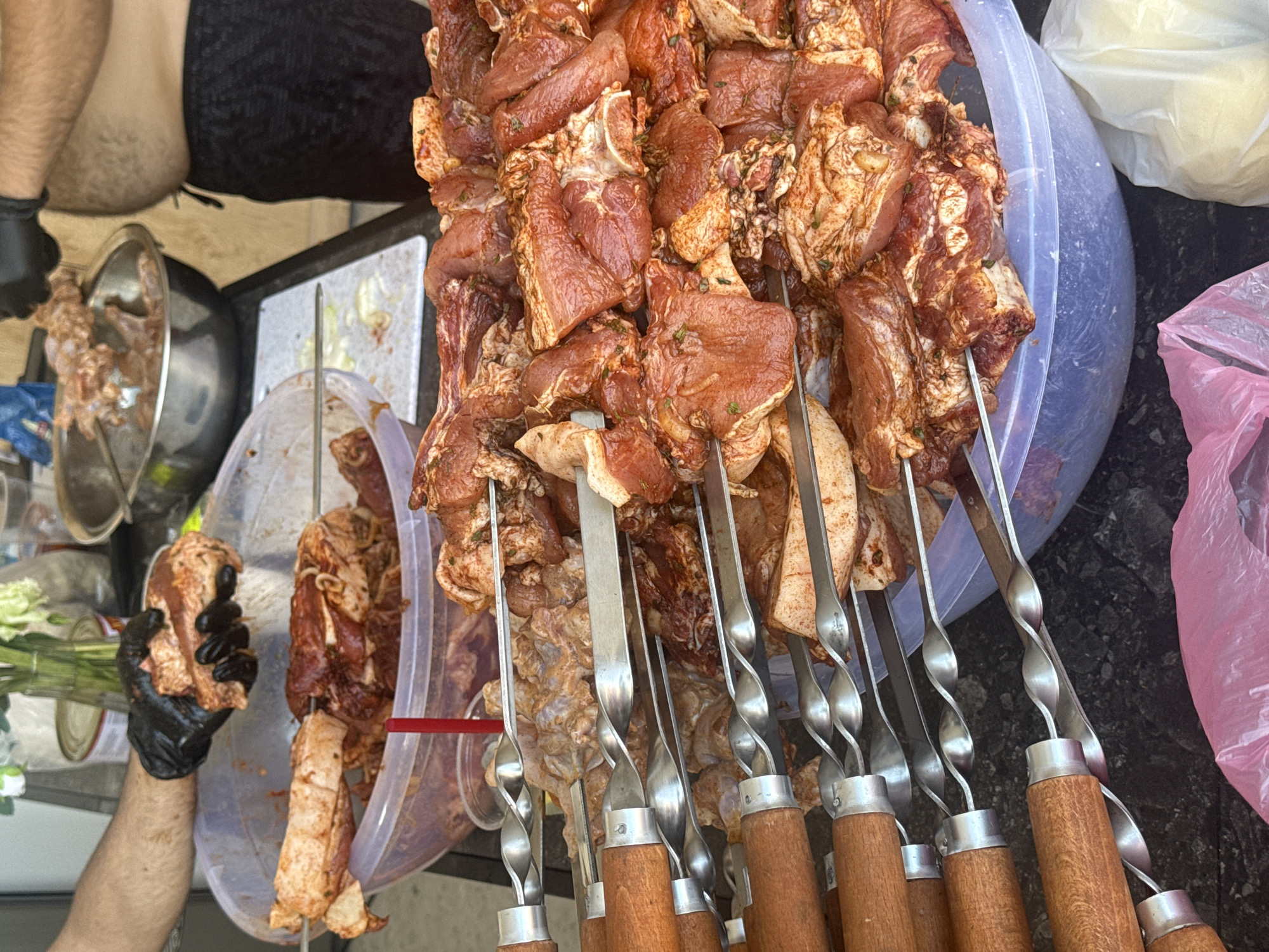 In Sasunik, Armenia, a man wearing black gloves is preparing meat skewers. He is holding a piece of marinated meat in his left hand, adding it to a large, clear plastic bowl filled with more marinated meat and skewers. In the background, another person is visible, also wearing black gloves and handling meat. The scene appears to be an outdoor preparation area, likely for a barbecue or event, with various bowls, containers, and cooking implements present. The meat is cut into bite-sized pieces and has a reddish-brown marinade with visible herbs. Several metal skewers, each twisted and holding pieces of meat, are arranged in the foreground and in the bowl. The overall impression is one of food preparation in progress.