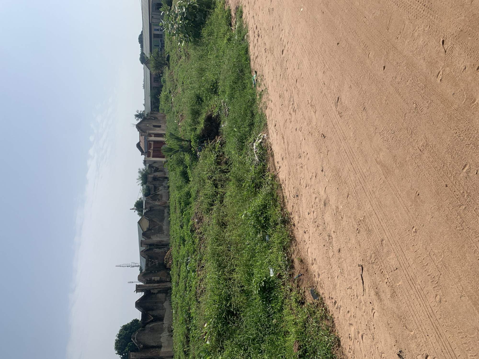 An eye-level, full outdoor shot captures a dirt road in the foreground, with tire tracks and footprints visible in the reddish-brown earth. To the left of the road, an overgrown, green grassy area is bordered by a broken, weathered concrete wall with jagged tops. Behind the wall, several partially constructed or derelict buildings are visible, some with corrugated metal roofs, along with scattered trees and communication towers in the distance against a hazy, pale blue sky. The overall scene appears to be an undeveloped or neglected area within a settlement.