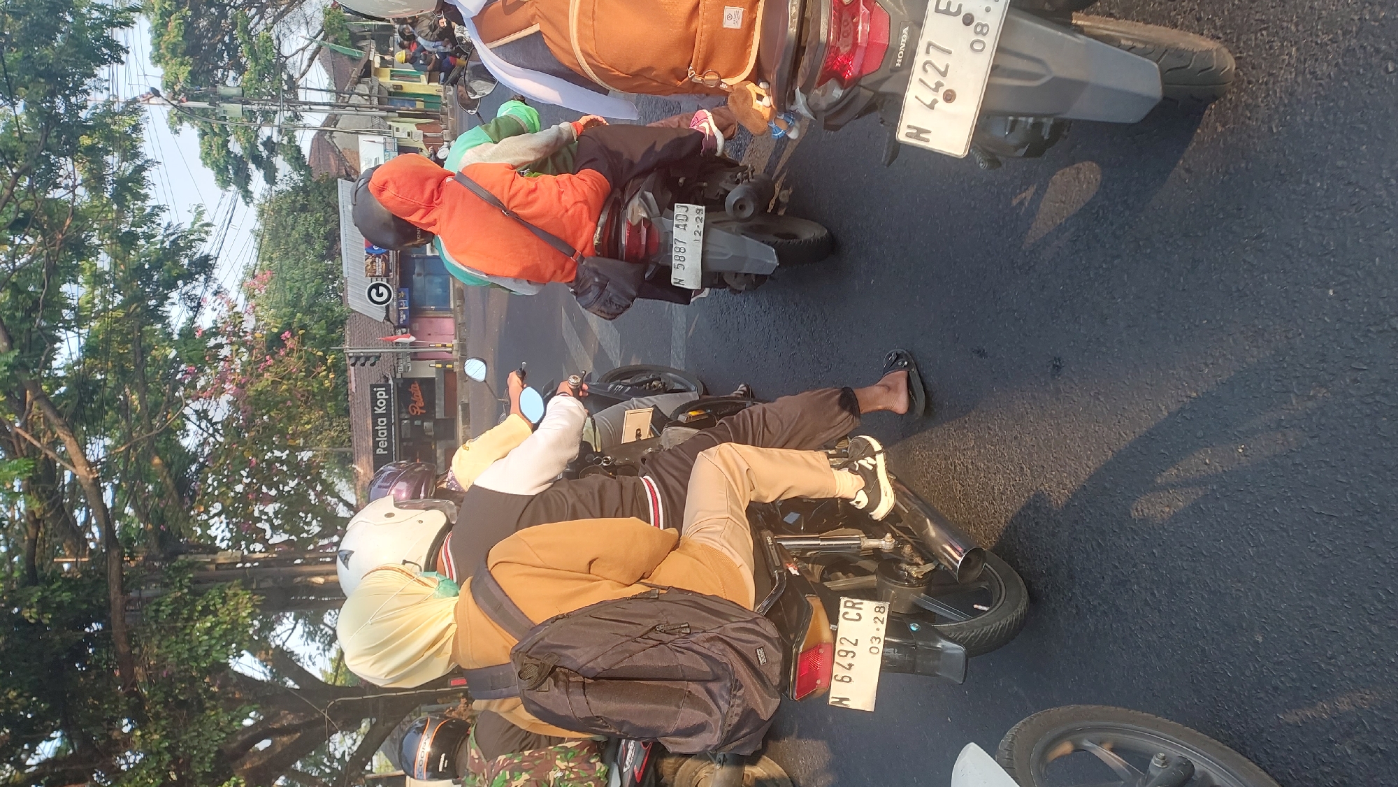 The image captures a street scene in Kesatrian, Indonesia, during daylight hours. The perspective is from behind several motorcycles moving along a paved road. On the left, a person wearing a yellow hijab and a tan jacket, carrying a black backpack, is seated on a motorcycle. They are wearing tan pants and black and white sneakers. The motorcycle's license plate reads "N 6492 CR 03-28". To their right, another motorcycle is visible with a rider wearing an orange hooded jacket and dark pants, carrying a dark bag. This motorcycle's license plate is "N 5887 ADJ 12-29". A third motorcycle, partially visible on the far right, has a rider with a brown backpack, and its license plate shows "N 4427 E 08-". Buildings and trees line the street, with one building prominently displaying "Pelata Kopi". The lighting suggests it is either morning or late afternoon, with shadows cast across the road. The atmosphere appears to be a typical urban traffic environment.