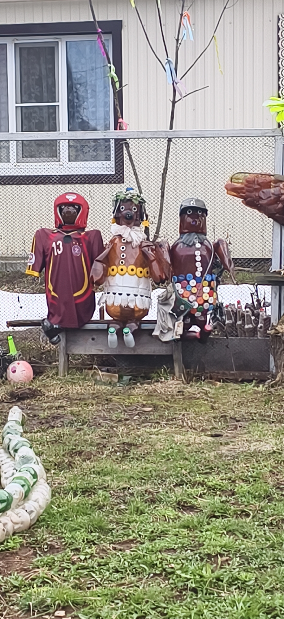 The image depicts three homemade sculptures sitting on a wooden bench in an outdoor setting, possibly a yard or garden. The sculptures are made from recycled materials. The first sculpture on the left wears a maroon and gold sports jersey with the number "13" on it, paired with a red headgear resembling a helmet. The middle sculpture is brown with a white-and-brown neckpiece and a head adorned with green accents. The sculpture on the right is brown, wearing a cap, and embellished with colorful bottle caps. The bench sits in front of a chain-link fence and a building with a window and light-colored siding. The foreground shows a grassy area with scattered debris. Based on the provided information, the location is Фоки, Russia.