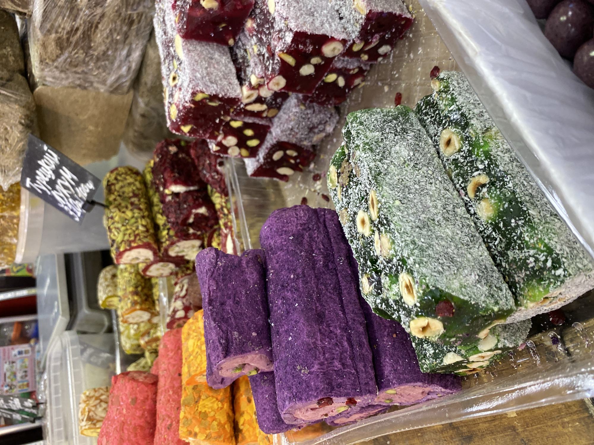 The image shows an assortment of Turkish Delight sweets in a plastic container, likely for sale. The sweets are arranged in rows, each row featuring a different flavor and color. From left to right, the rows include pink, orange, purple, green, yellow, and dark red pieces. The dark red pieces are studded with pistachios, while the green pieces contain hazelnuts. The sweets are dusted with a powdery substance, likely confectioner's sugar. A small sign with Cyrillic text is visible to the left of the sweets. The location is likely a market or store in the city of Chaikovsky, Russia.