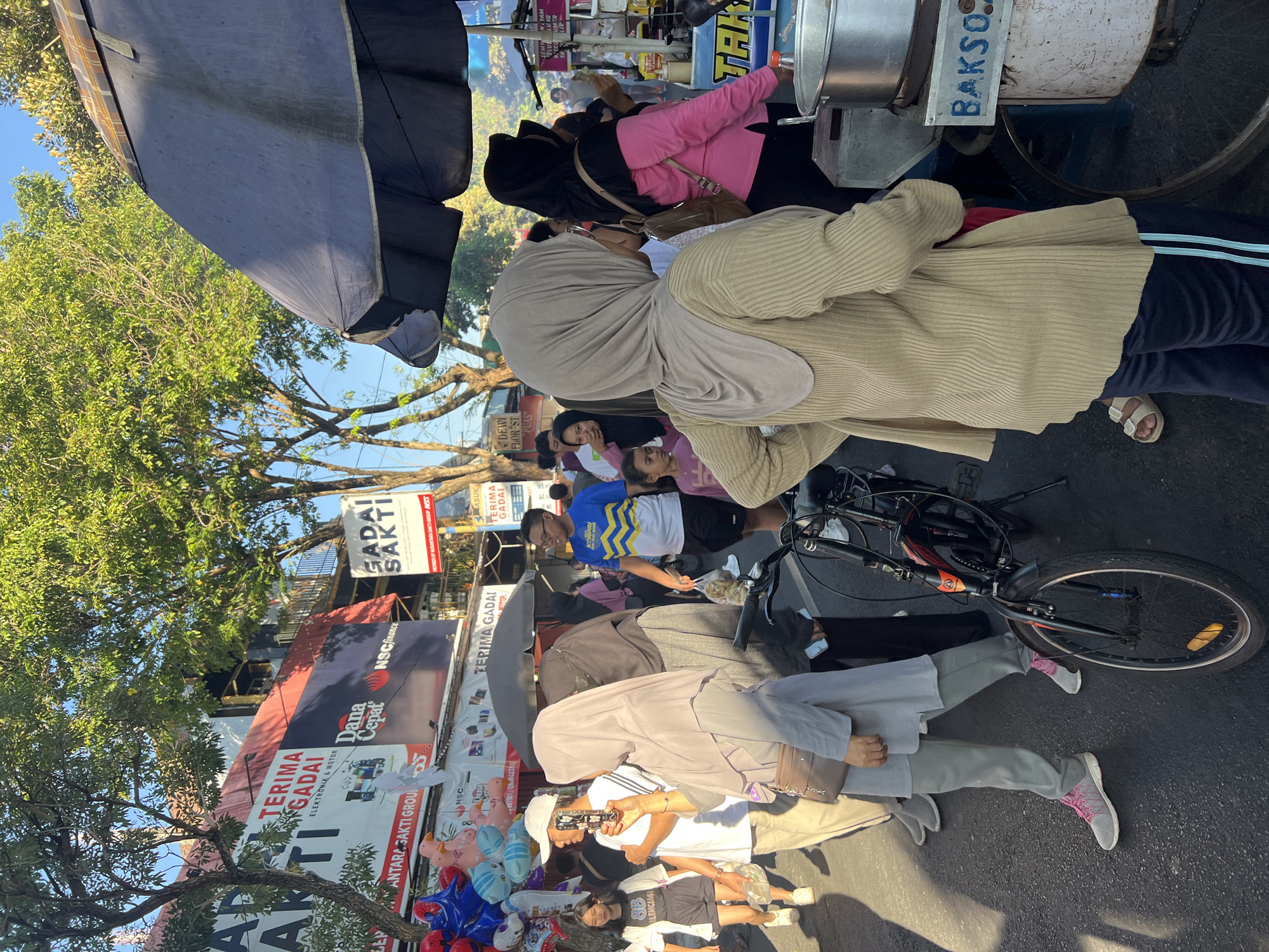 The image captures a bustling street scene in Kecamatan Pati, Indonesia, bathed in the warm glow of a late afternoon sun. The foreground is dominated by a sidewalk populated with pedestrians and vendors. A woman in a light brown hijab and a beige ribbed cardigan is looking away from the camera, her left hand resting on her hip. Beside her, a bicycle with a red frame is parked. To the left of the bicycle, a person wearing a grey hijab and grey pants with pink and grey sneakers is seen from the waist down.
Further back, a vendor's stall with a large blue and white sign that reads "BAKSO" is visible, featuring a metal pot. In the background, more people are walking along the street, and various buildings with signs can be observed. One prominent sign displays "TERIMA GADAI" (Accepts Pawn) and "DANA CEPAT" (Fast Cash). Another sign reads "GADAI SAKTI". Trees with lush green foliage line the street, casting shadows on the pavement. The overall atmosphere suggests a lively commercial area with everyday street life unfolding.