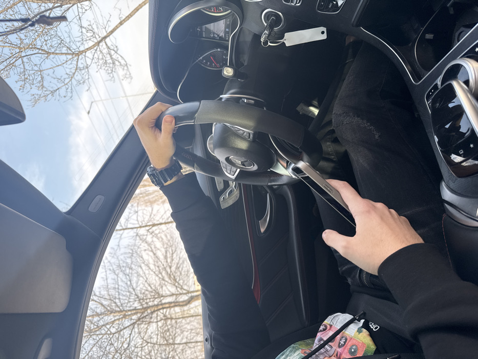 The image depicts the interior of a modern Mercedes-Benz vehicle, viewed from a slightly elevated position behind the driver. A person, presumed to be the driver, is shown with their right arm and hand gripping the top of the black multi-function steering wheel. A dark digital watch is visible on their right wrist, and their arm is covered by a black long-sleeved garment. To the lower left, another person's left hand holds a silver-colored smartphone upright. This individual is wearing black distressed jeans with visible rips and a multi-colored patterned hoodie.
The car's interior features a black dashboard, door panels with a subtle red accent line, and chrome trim. The instrument cluster shows analog dials and a digital display. Car keys with a blank white key tag hang from the ignition or a dashboard hook. The center console contains a glossy control panel with buttons and a rotary selector. An "AIRBAG" label is visible on the A-pillar above the driver's side.
Through the vehicle's windows, a clear blue sky is visible. Leafless deciduous trees line the background, and a utility pole with overhead power lines is discernable in the distance to the right. The general environment appears to be an urban or suburban setting. No specific landmarks or unique architectural features are present to definitively confirm the location as Sofia, Bulgaria.