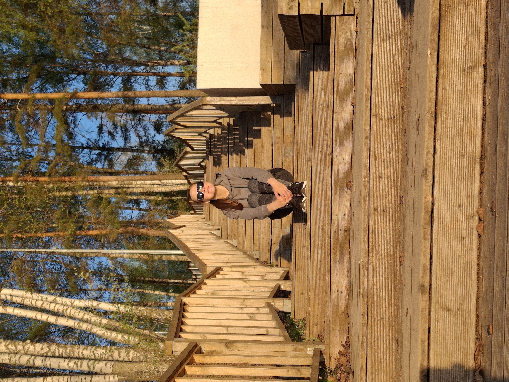The image shows a woman wearing sunglasses, a gray hooded sweatshirt, and dark pants, sitting on a wooden platform in what appears to be an outdoor recreational area. She is positioned at the top of a long wooden staircase. The staircase leads down towards birch trees and other foliage. Behind her, there is a wooden structure with benches. The lighting suggests it might be late afternoon or early evening. Based on the location metadata, this scene is likely in Chaikovsky, Russia.