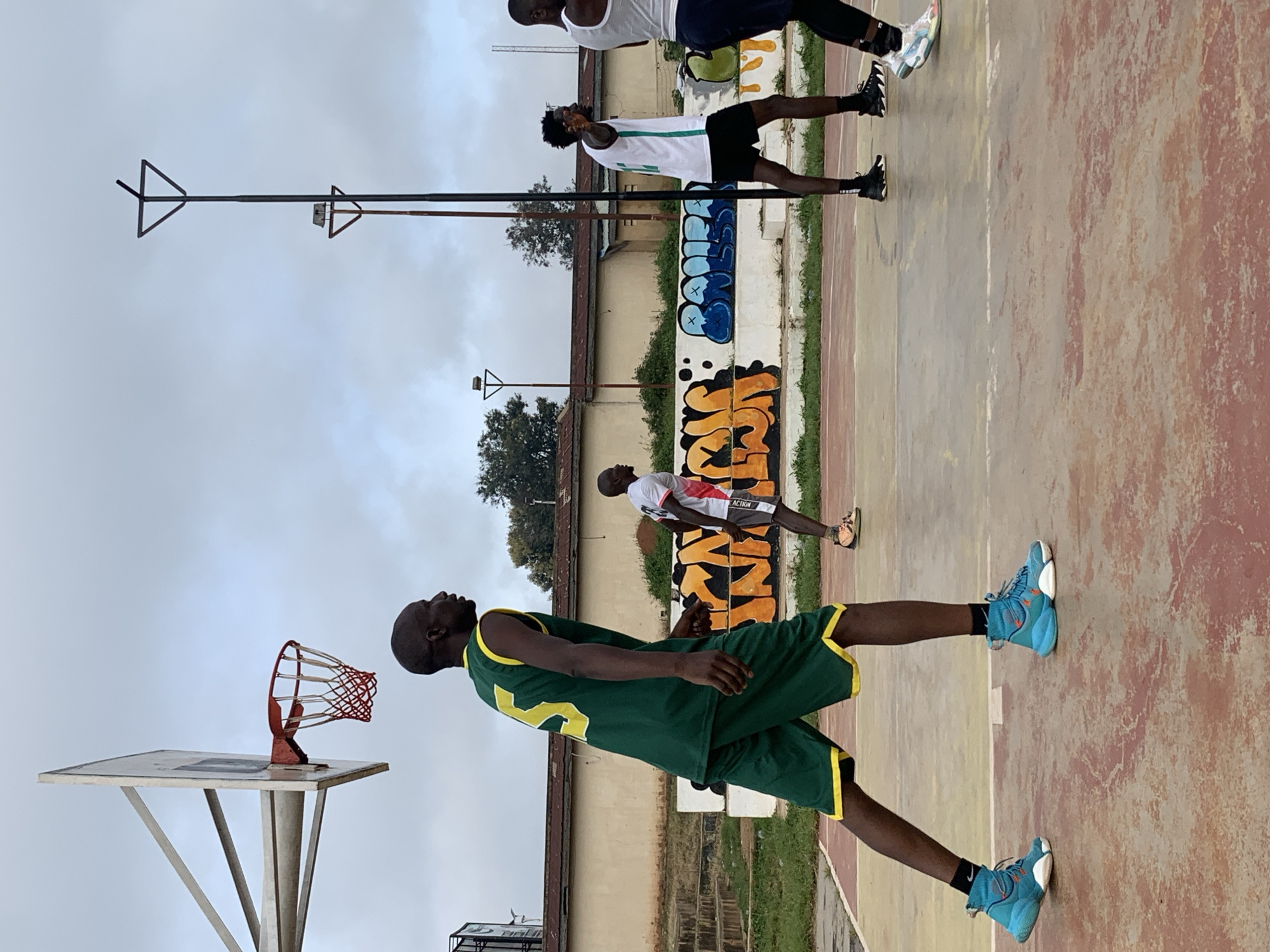 The image depicts a basketball court scene. In the foreground, a young Black man wearing a green basketball jersey with yellow trim and the number 5 on the back, and matching green shorts with yellow trim, stands in a defensive stance. He is wearing bright blue basketball shoes. Behind him, to his right, another young Black man in a white basketball jersey with green trim and dark shorts is visible, facing away from the camera. Further in the background, a third young Black man in a white tank top and dark shorts is partially visible on the right side of the frame. The basketball court has a reddish-brown and yellow surface with white lines. A basketball hoop with a red rim and net is visible on the left. Behind the court, there is a low wall with colorful graffiti. The environment suggests an outdoor court, possibly in a neighborhood setting, with some greenery and buildings in the background. The sky is overcast. The image appears to be taken in Jos, Nigeria.