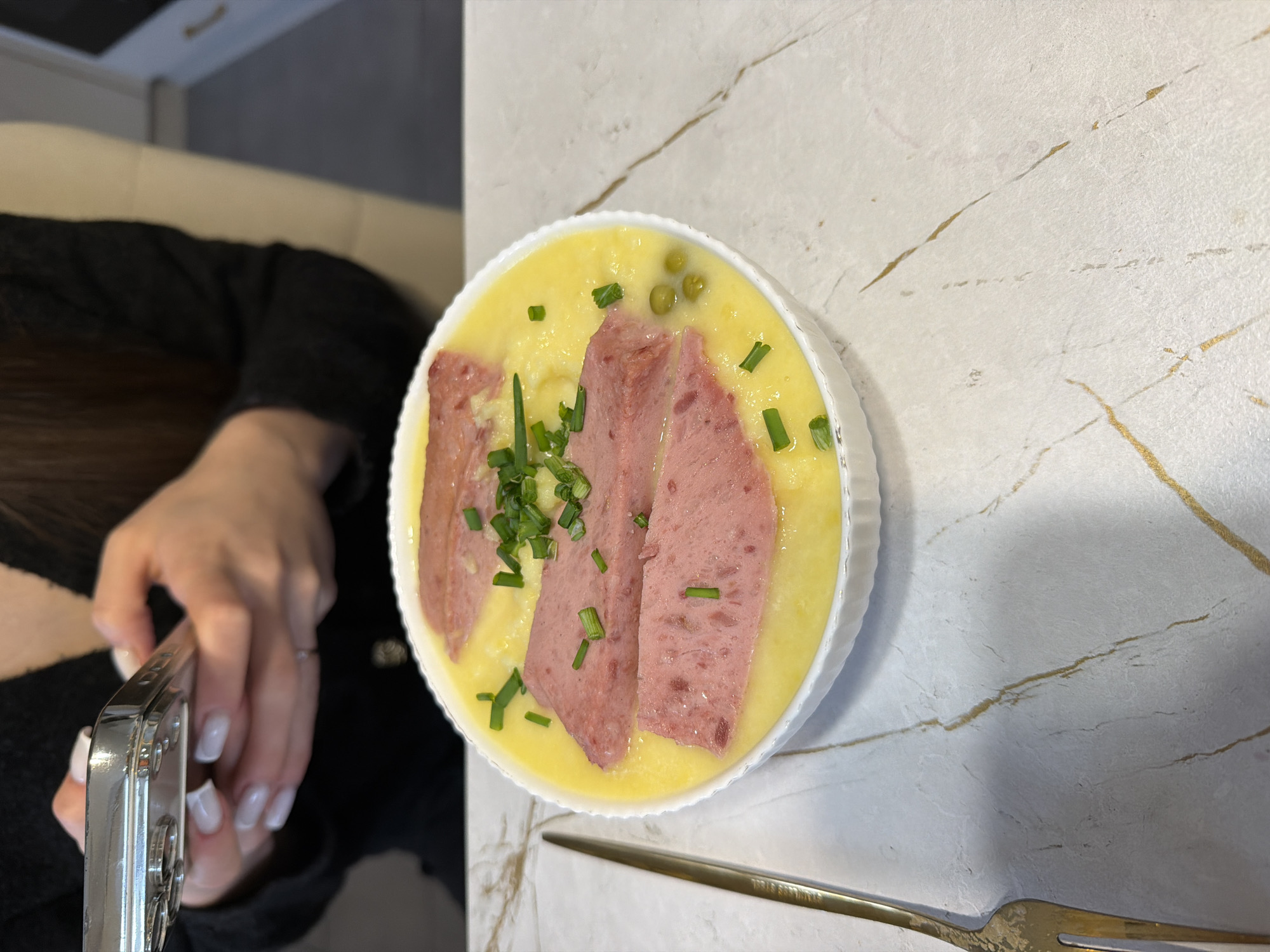 A close-up, slightly angled, high-angle shot shows a white ceramic dish containing a light yellow puree topped with sliced meat, green peas, and chopped chives. The dish rests on a white marble-patterned countertop with golden veins. A gold fork sits diagonally across the lower right corner of the frame. In the lower-left, part of a person's arm and hand holding a silver phone is visible. The person has manicured nails with white polish and a thin silver ring on their finger. The person wears a dark-colored long-sleeved shirt. The scene takes place indoors. The location is not identifiable in this image as being Chaikovsky, Russia.