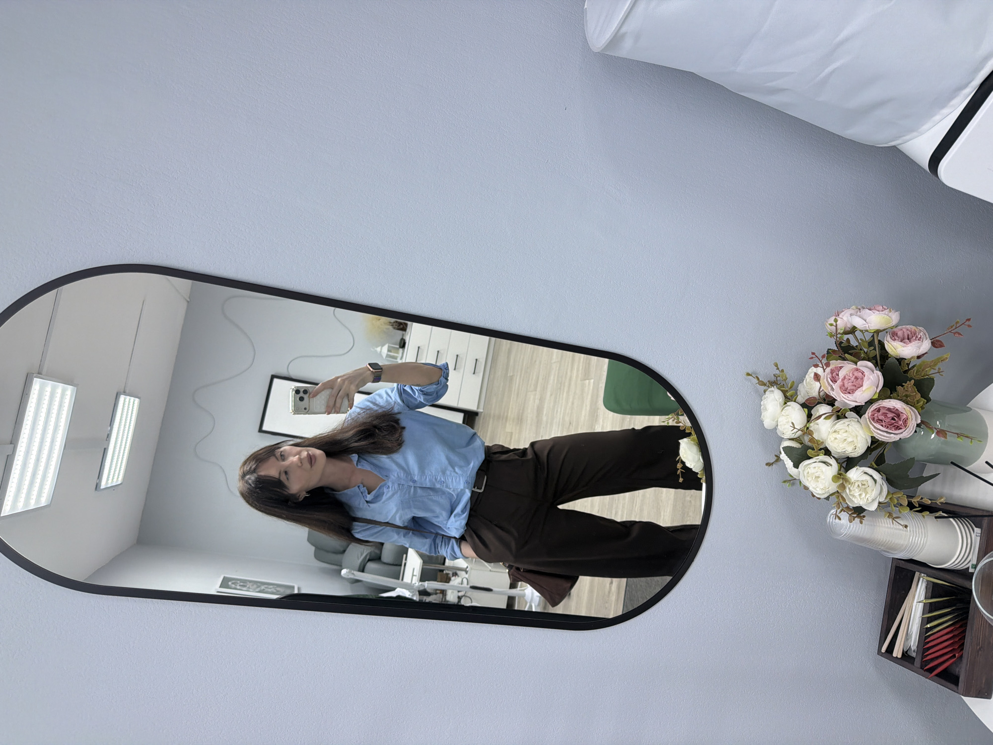 A woman is taking a selfie in a mirror. The mirror is oval with a black frame. The woman is wearing a light blue shirt and dark brown pants. She is holding a smartphone in her right hand. The background of the mirror shows a room with dental equipment. A vase of flowers and a container with cups and sticks are located on the wall near the mirror. The location is Perm, Russia, as indicated by the reflection of dental equipment which is commonly used in dental offices and clinics there.